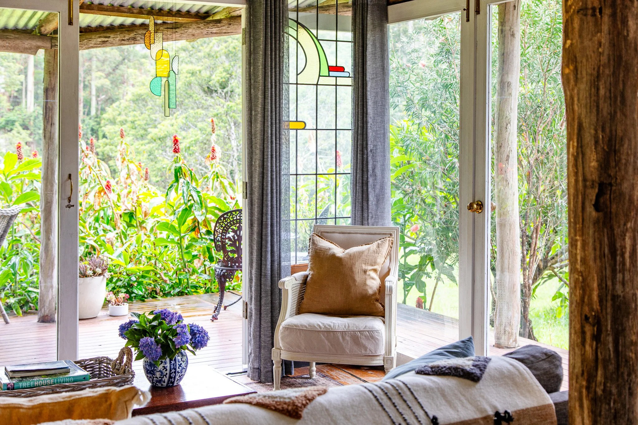 A cozy living room with open glass doors leading to a deck with lush garden outside. There is a white armchair with a brown cushion, a small table with a vase of purple flowers, and a sofa with a blanket. Decor includes stained glass hanging art and 