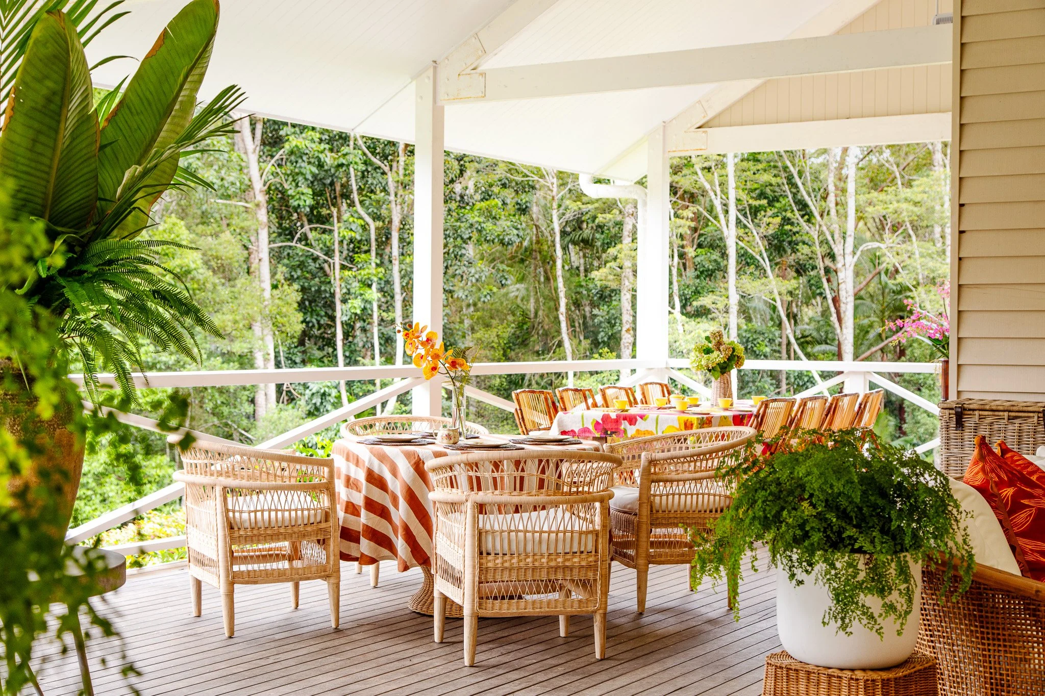 A bright outdoor porch with dining table, surrounded by woven chairs, decorated with colorful tablecloths, flowers, and lush green trees in the background.