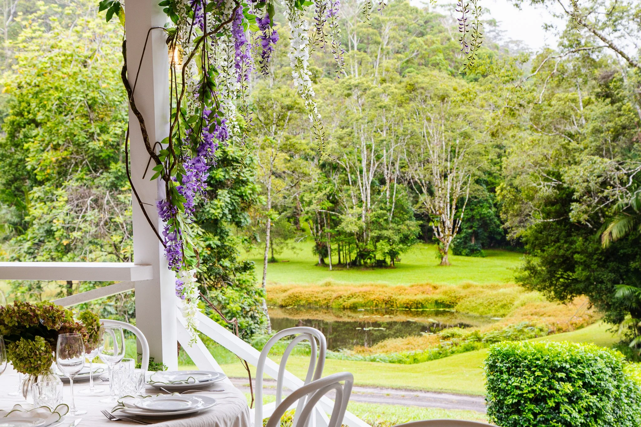 A table set for a meal outdoors on a porch overlooking a lush green landscape with trees, a pond, and grass. Purple and white wisteria flowers dangle from above.