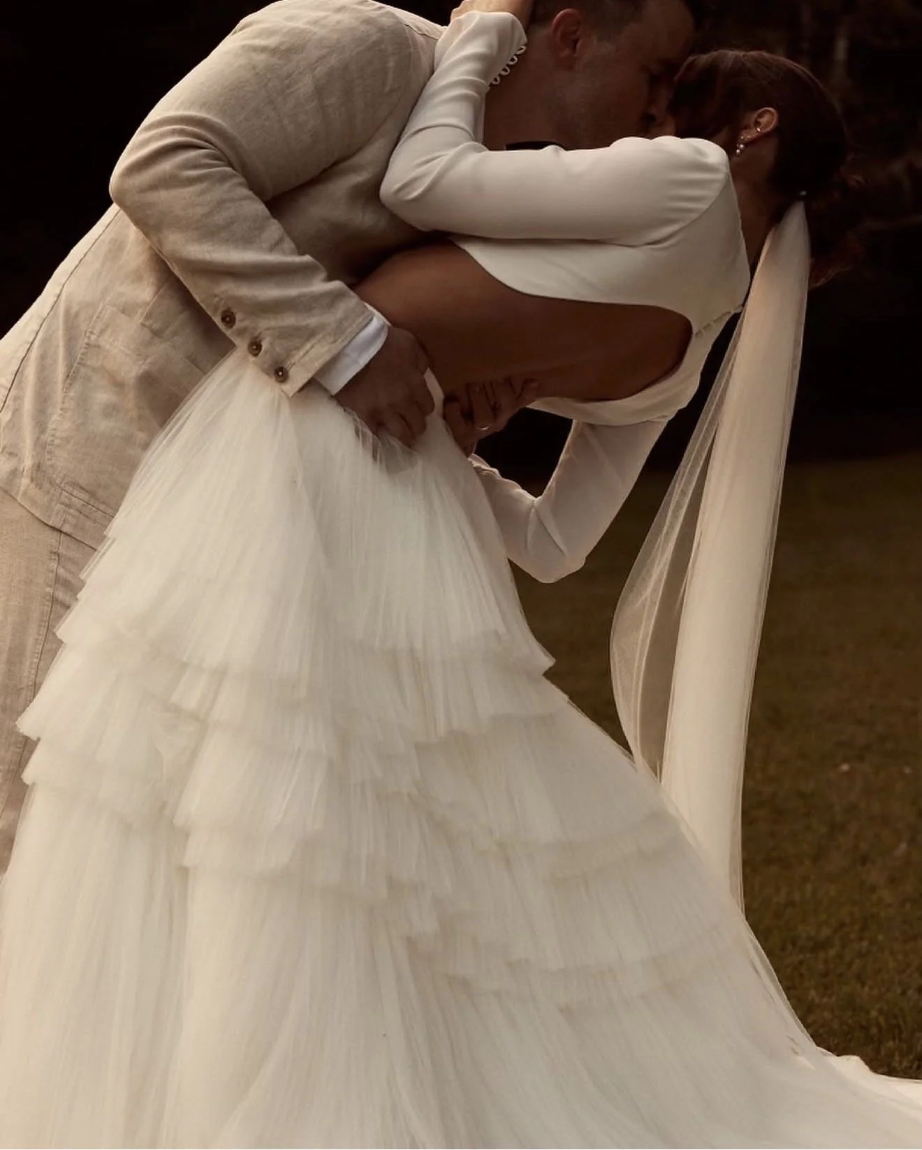A couple dressed in wedding attire sharing a kiss, with the groom in a light-colored suit and the bride in a white wedding gown with a layered skirt and long veil.