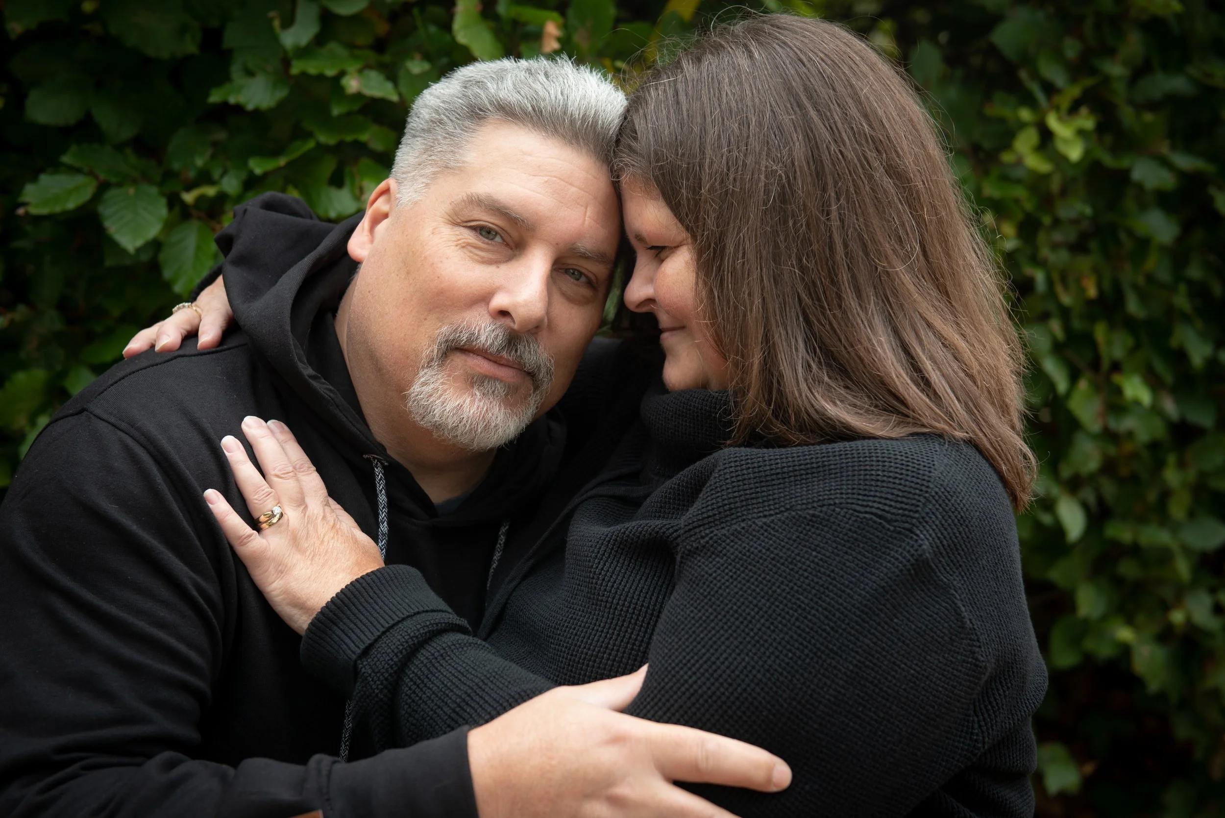 A couple hugging outdoors with green foliage in the background.