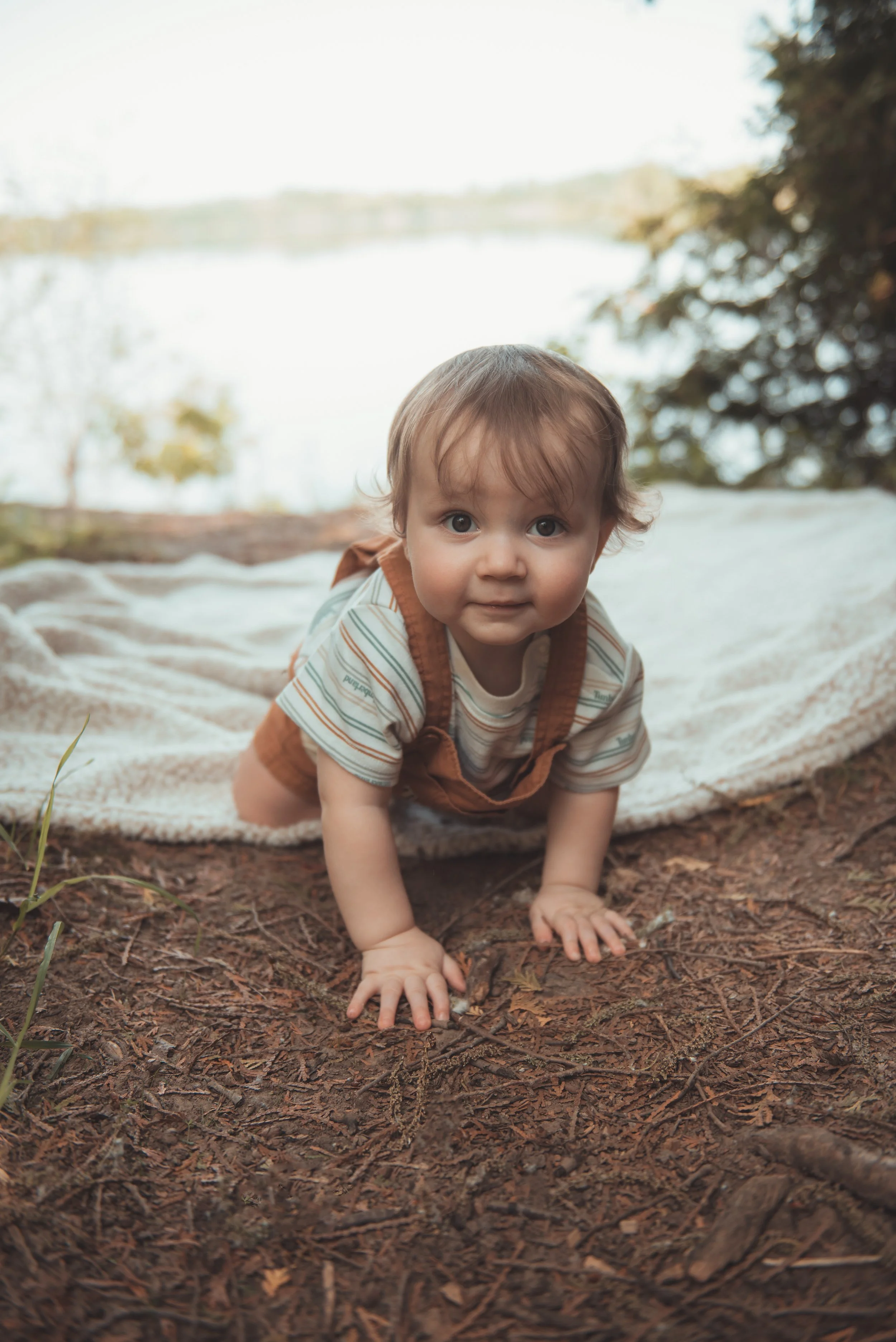 Baby crawling on a forest floor with a lake and trees in the background.