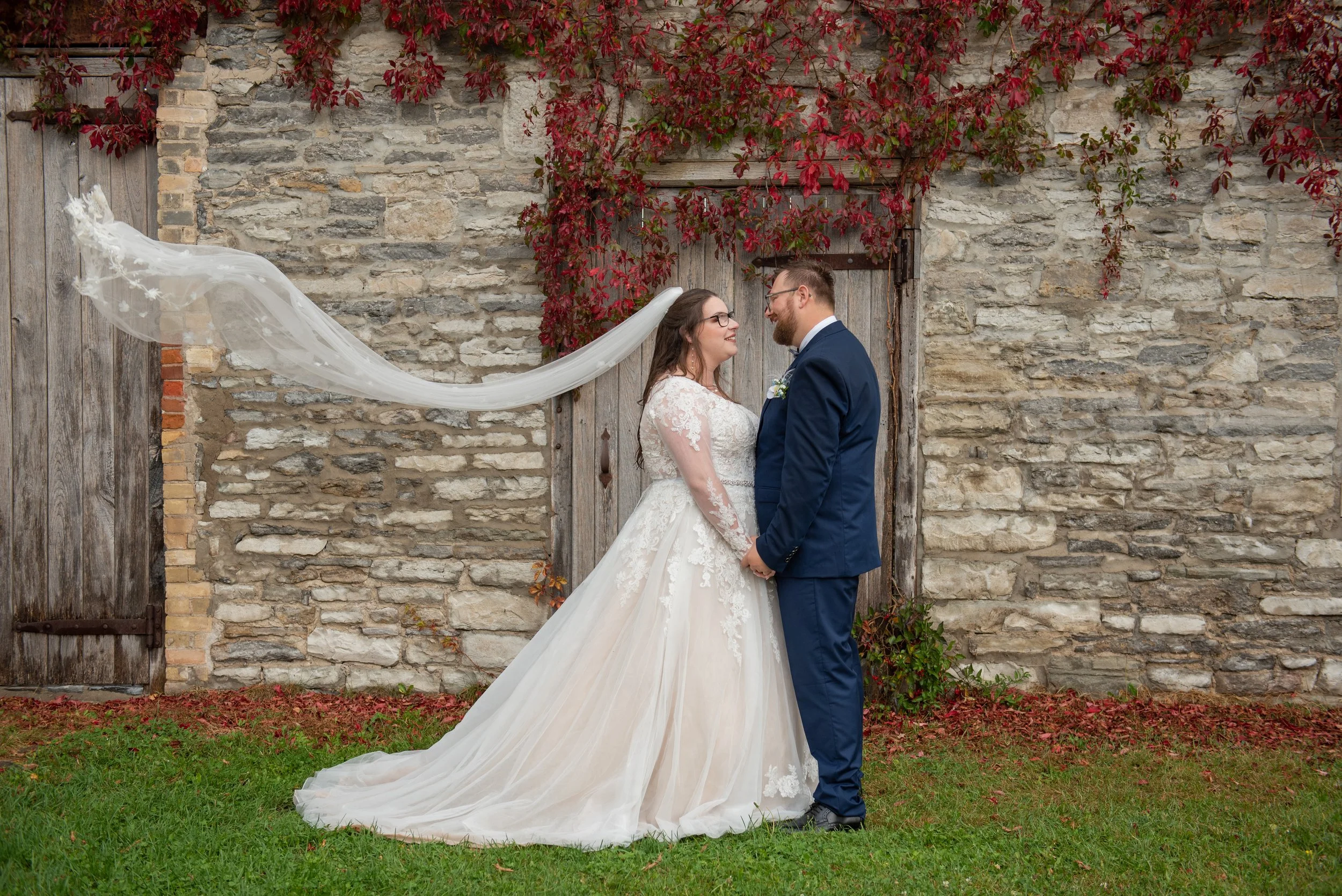 A bride and groom stand facing each other outdoors in front of a stone wall with climbing red leaves. The bride wears a white lace wedding gown with a long train and veil, while the groom wears a navy blue suit. They hold hands and look into each other's eyes.