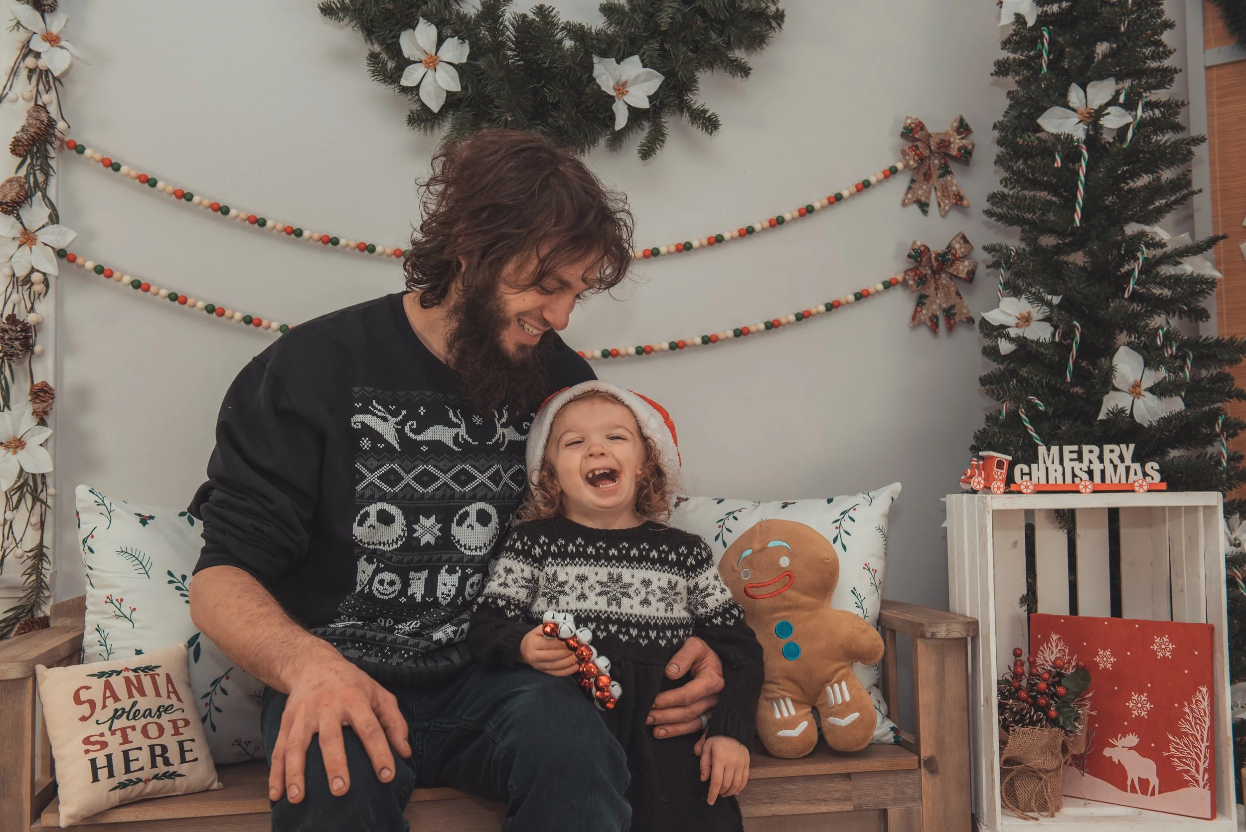 A man and a young girl sitting on a bench in Christmas decorations, laughing. The girl is holding jingle bells and wearing a Santa hat. The man has a beard and is wearing a Christmas sweater. There is a gingerbread cookie plush and a big Christmas tree decorated with candy canes, flowers, and ornaments behind them. Christmas-themed pillows and signs are also present.