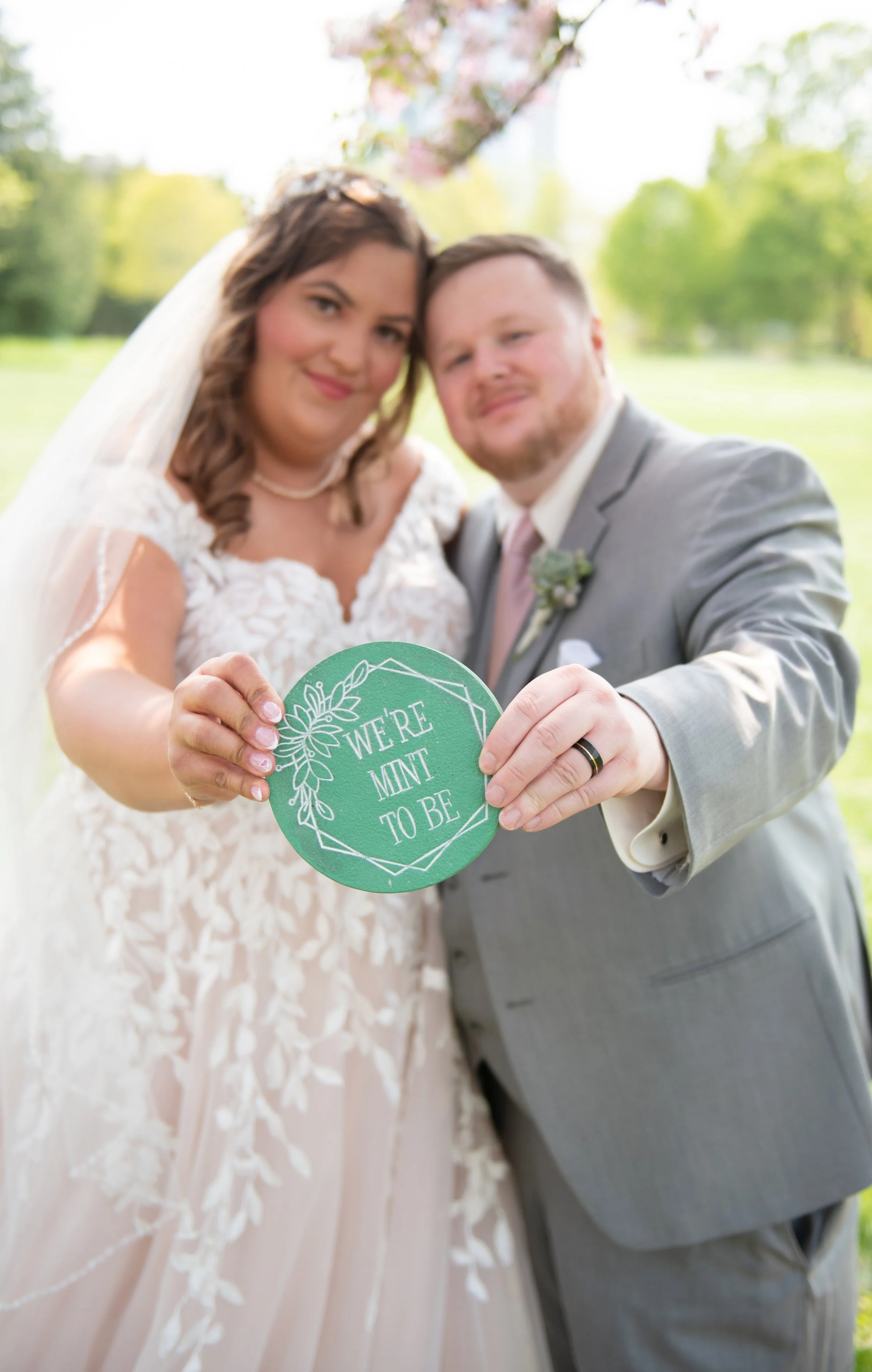 A newlywed couple holding a green circular sign that says 'We're Mint to Be' at their wedding outdoors.