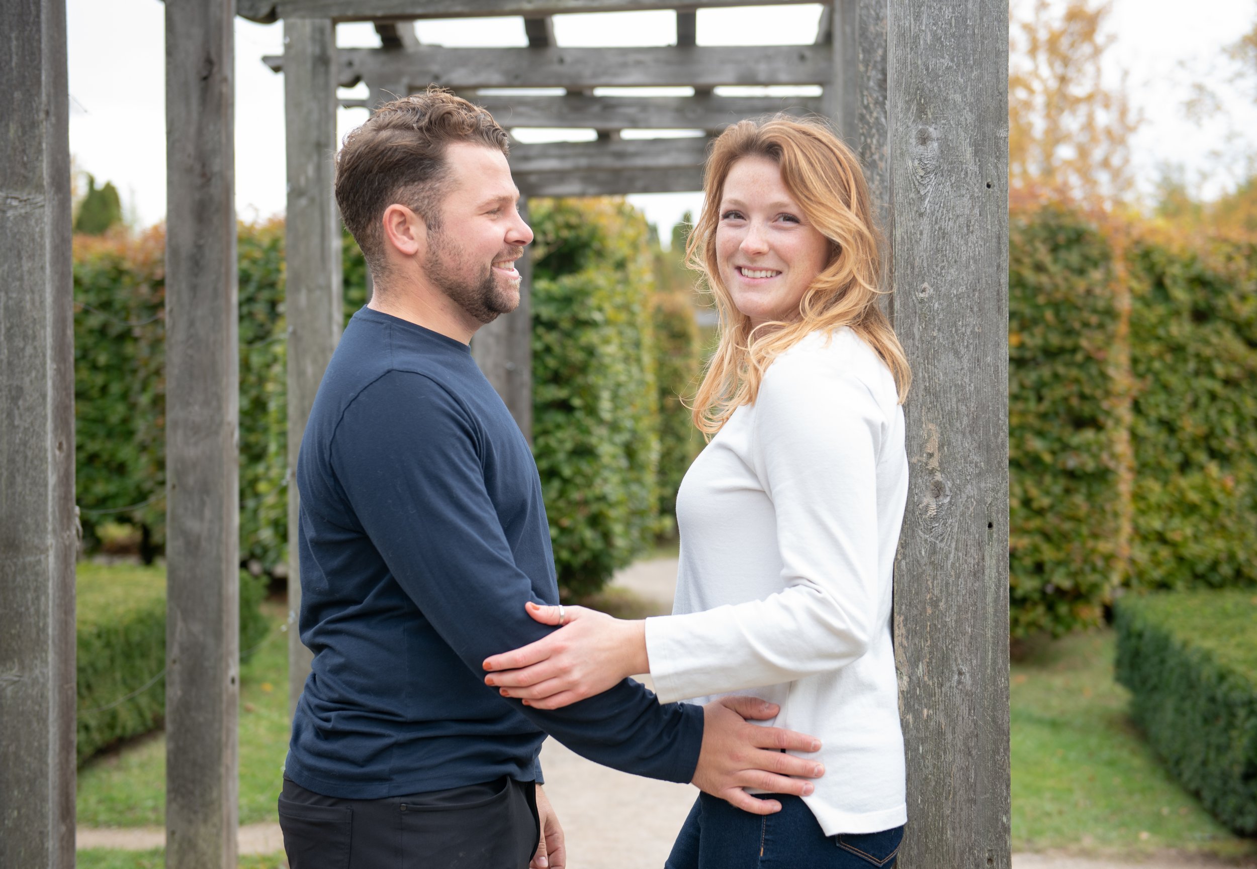 A smiling man and woman standing outdoors, facing each other and holding hands, with trees and a wooden pergola in the background.