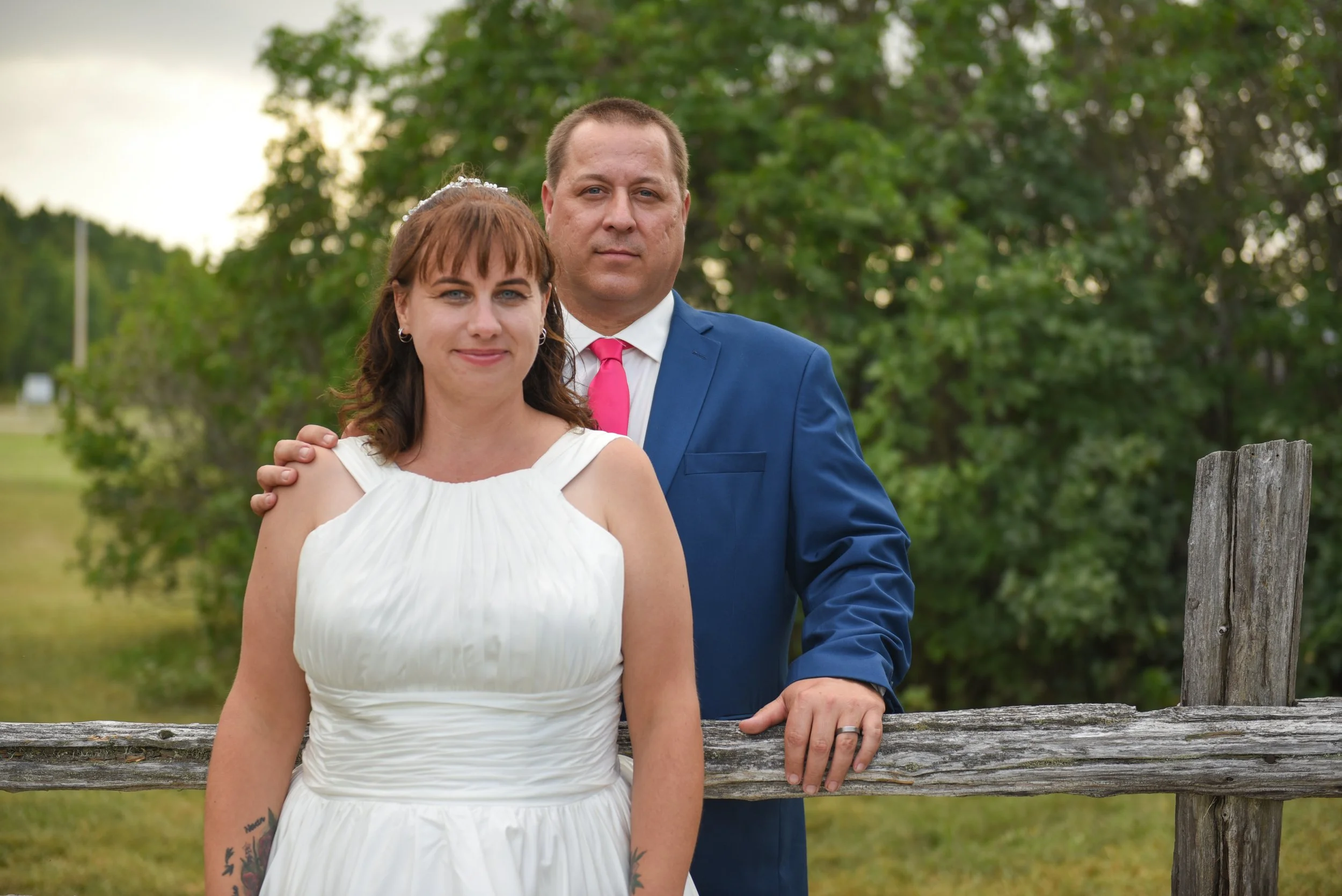 A couple dressed in formal attire, standing outdoors near a wooden fence with green trees in the background. The woman is wearing a white dress and has tattoos on her left arm. The man is wearing a blue suit with a pink tie, and has his right hand resting on the fence behind the woman.