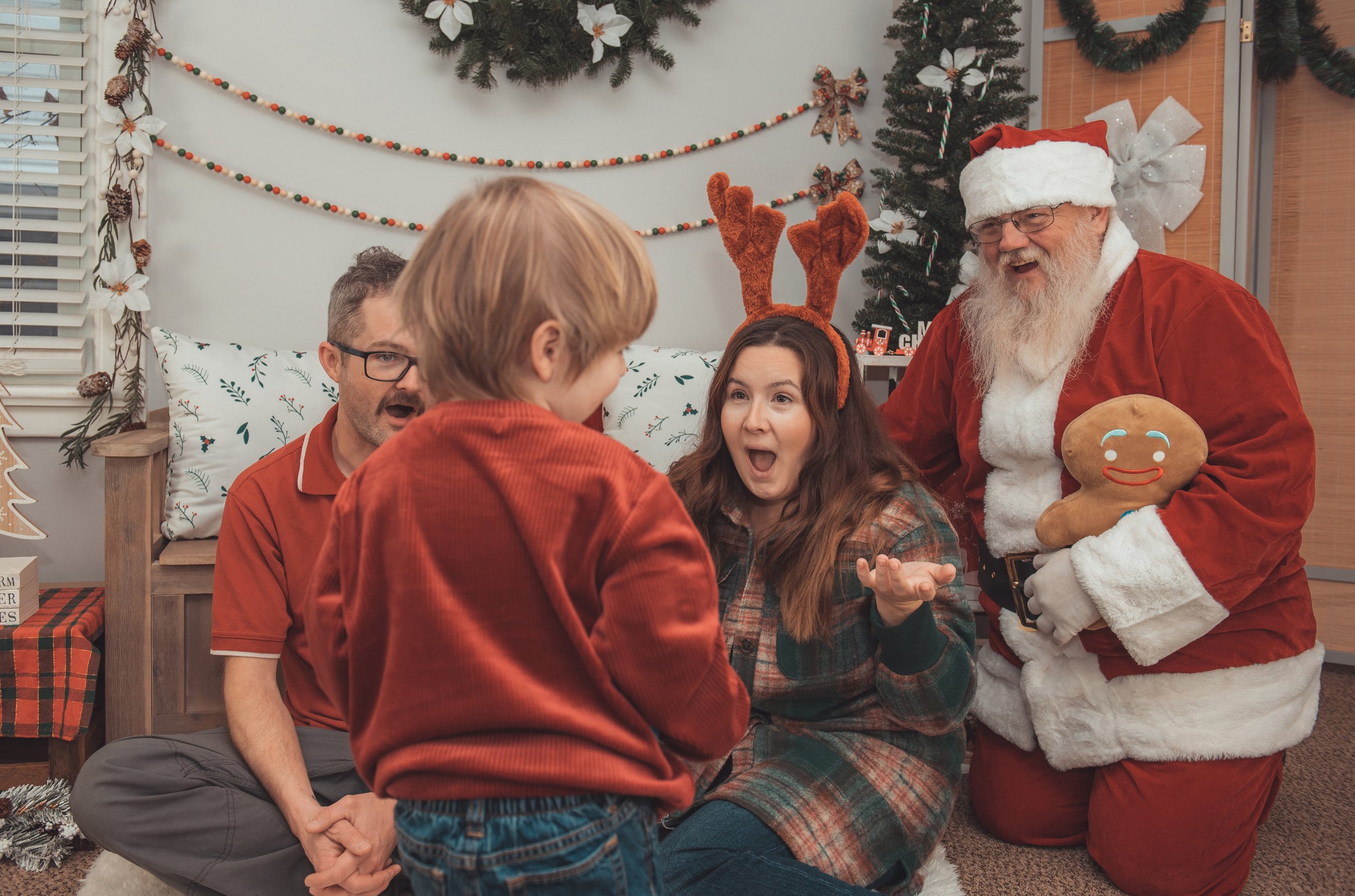 Four people, including a man dressed as Santa Claus holding a gingerbread man plush, are happily interacting with a young boy wearing reindeer antlers. They are in a room decorated with Christmas ornaments, garlands, and a tree, celebrating the holiday.