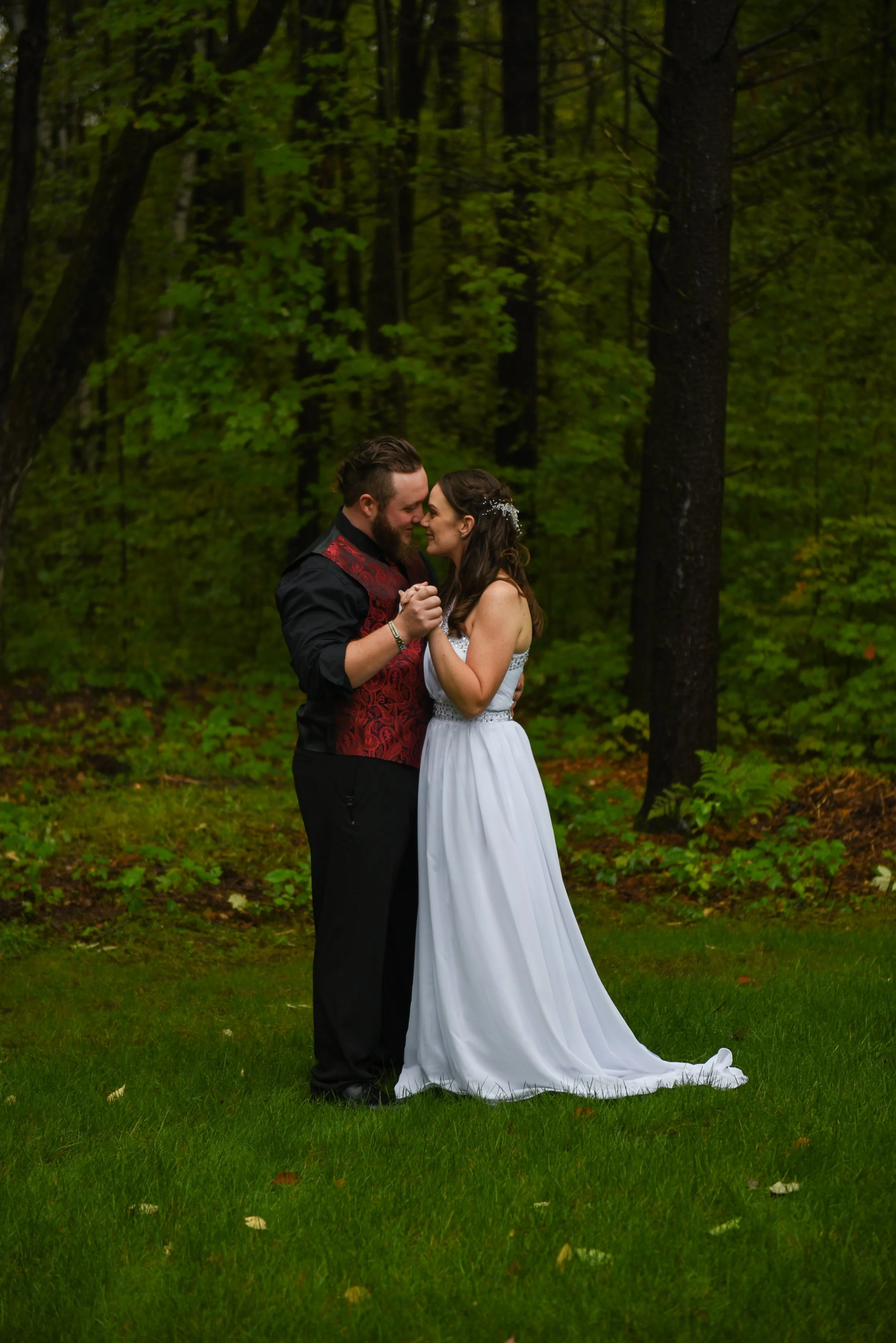 A couple in wedding attire standing close in a green forest, holding hands and touching foreheads.