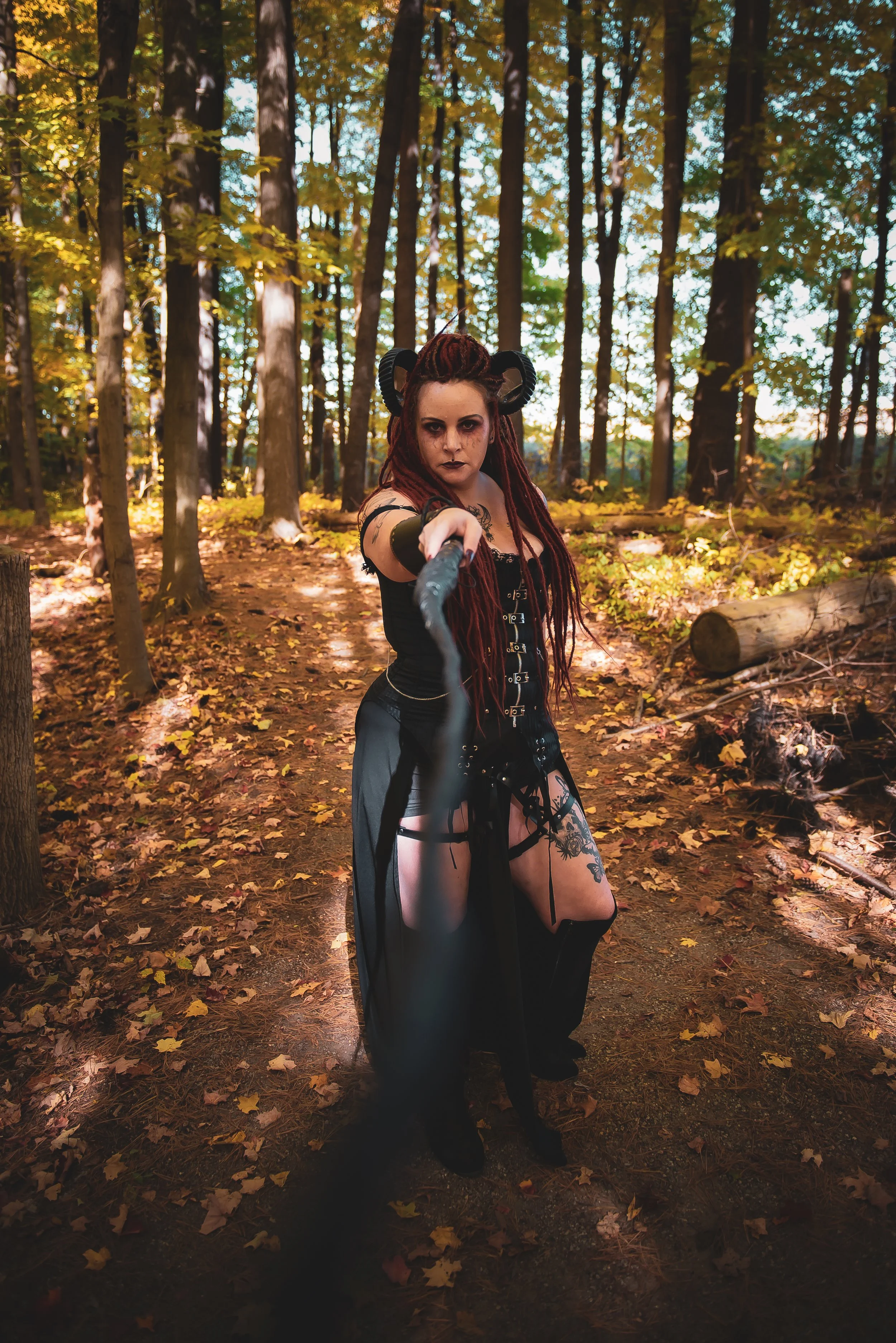 A woman dressed in dark, Gothic-style clothing with horns on her head, standing in a wooded forest during daytime, holding a whip towards the camera.