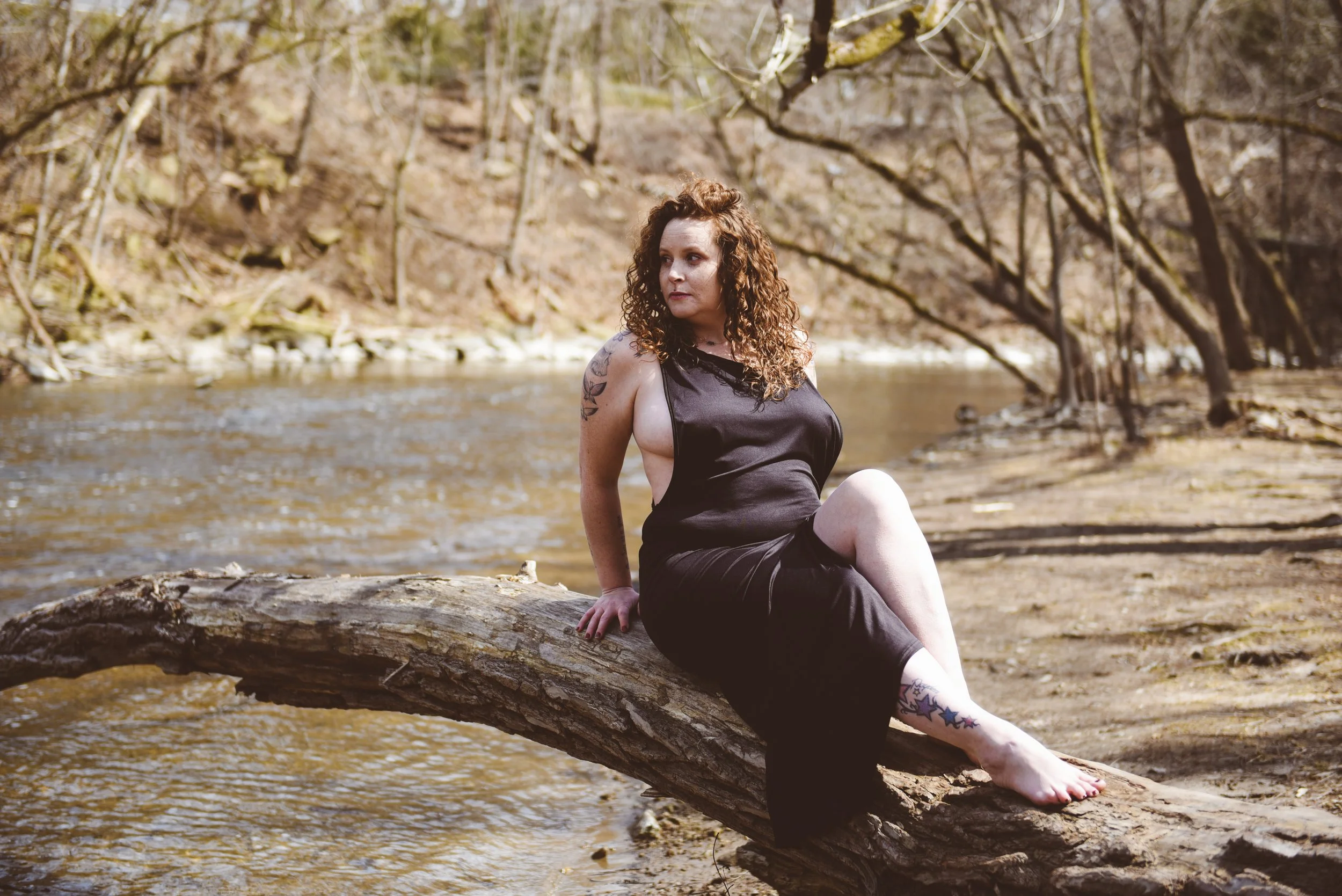 A woman with curly hair sitting on a fallen tree trunk by a river, surrounded by trees with bare branches in a forest.