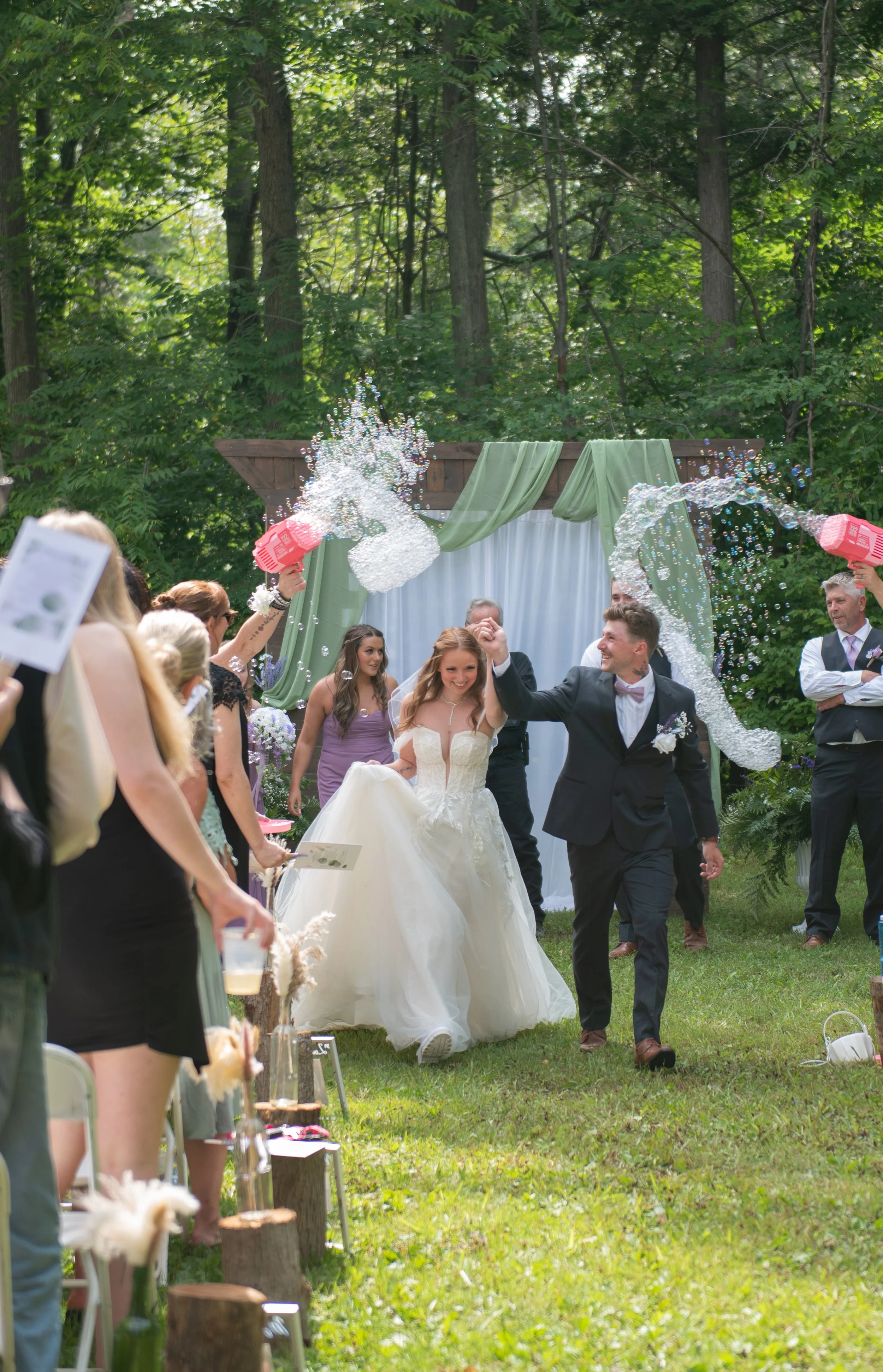 A newlywed couple walking hand in hand through an outdoor wedding ceremony, surrounded by friends and family, with soap bubbles being blown around them, in a lush green forest setting.