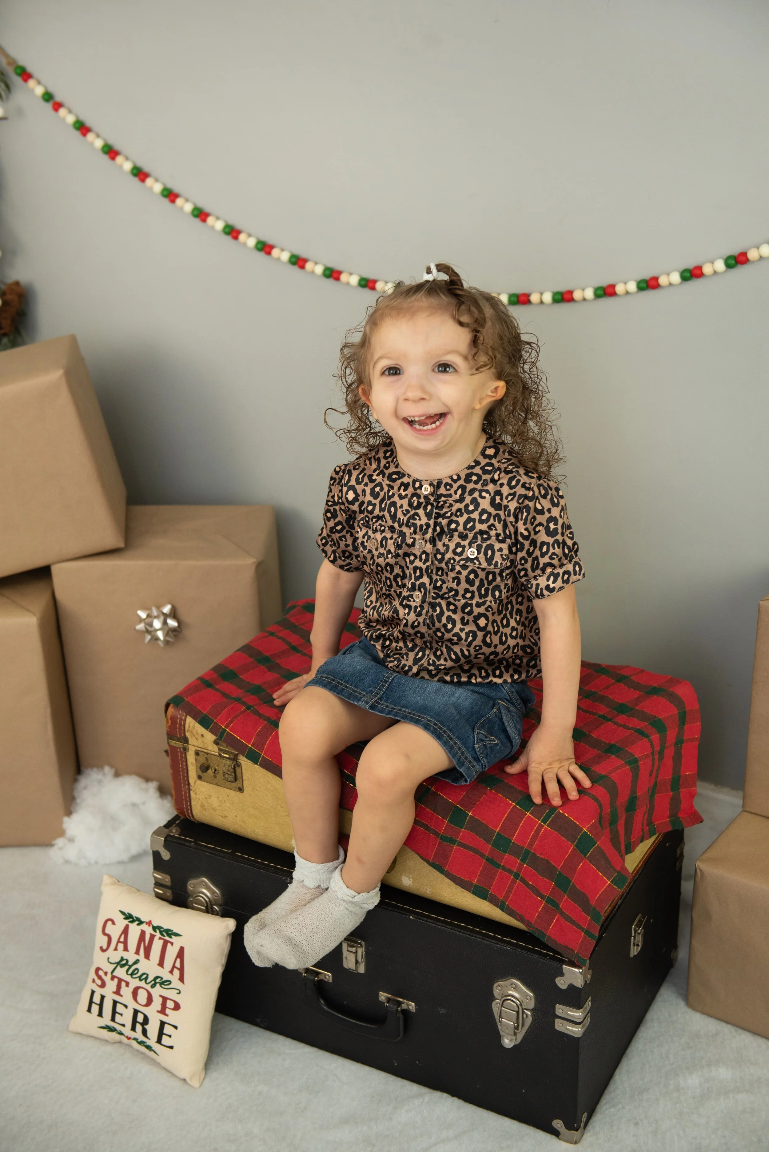 A smiling young girl with curly hair sitting on top of a vintage black suitcase in a festive holiday setting, wearing a leopard print shirt and denim skirt, surrounded by wrapped gifts and Christmas decorations.