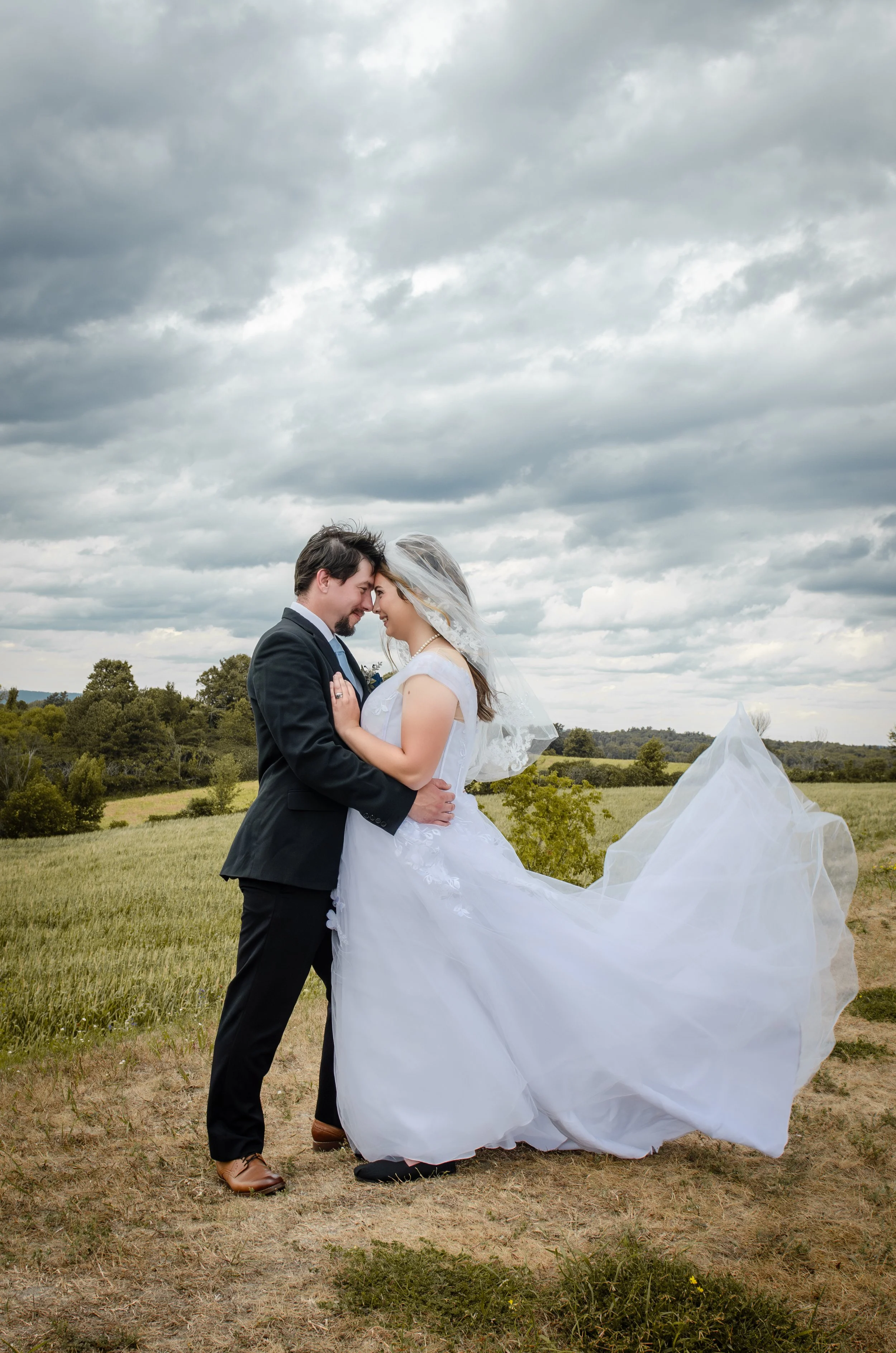 A bride and groom in an embrace outdoors, with rolling hills and scattered trees under a cloudy sky.