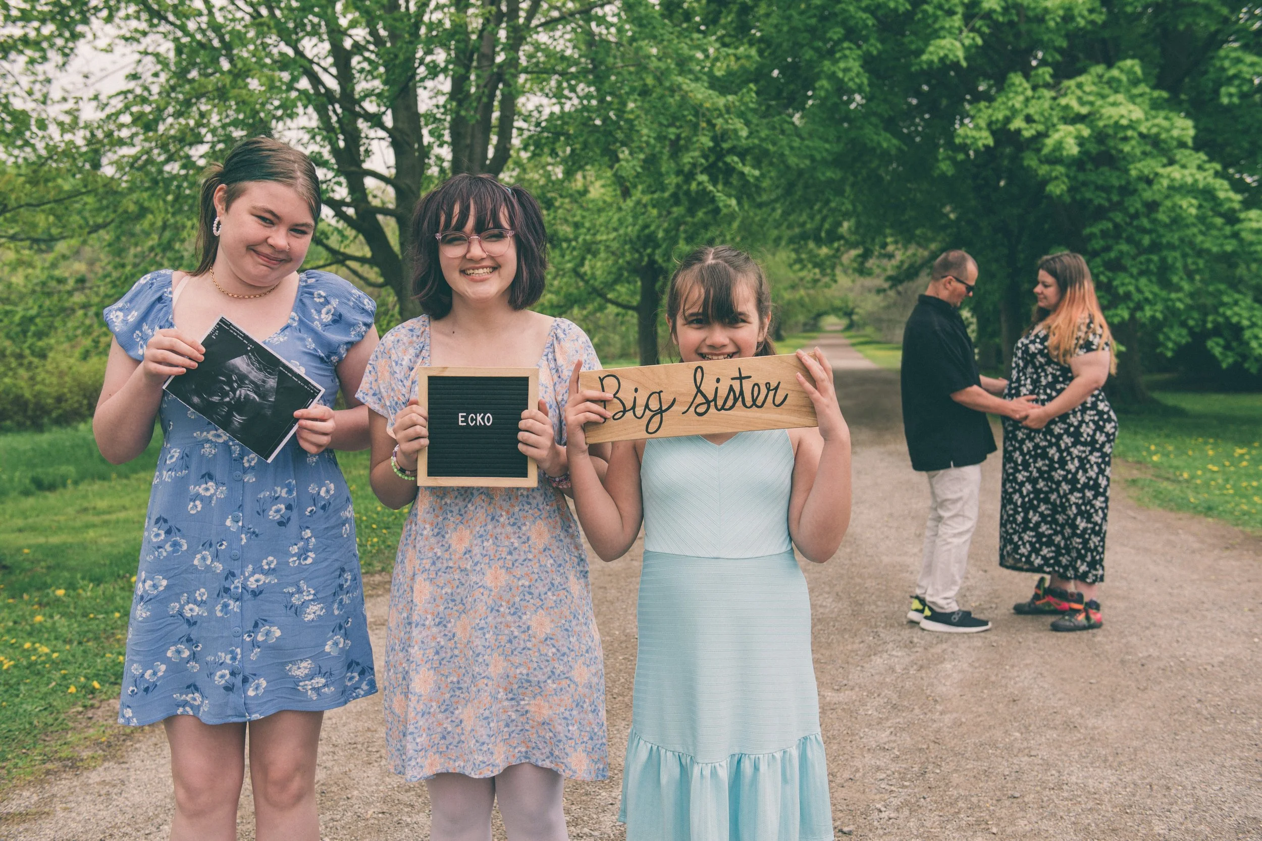Three young women standing on a park path, smiling. The woman on the left holds an ultrasound picture, the woman in the middle holds a sign saying 'Ecko', and the woman on the right holds a sign saying 'Big Sister'. In the background, a man and a woman are seen holding hands and facing each other.