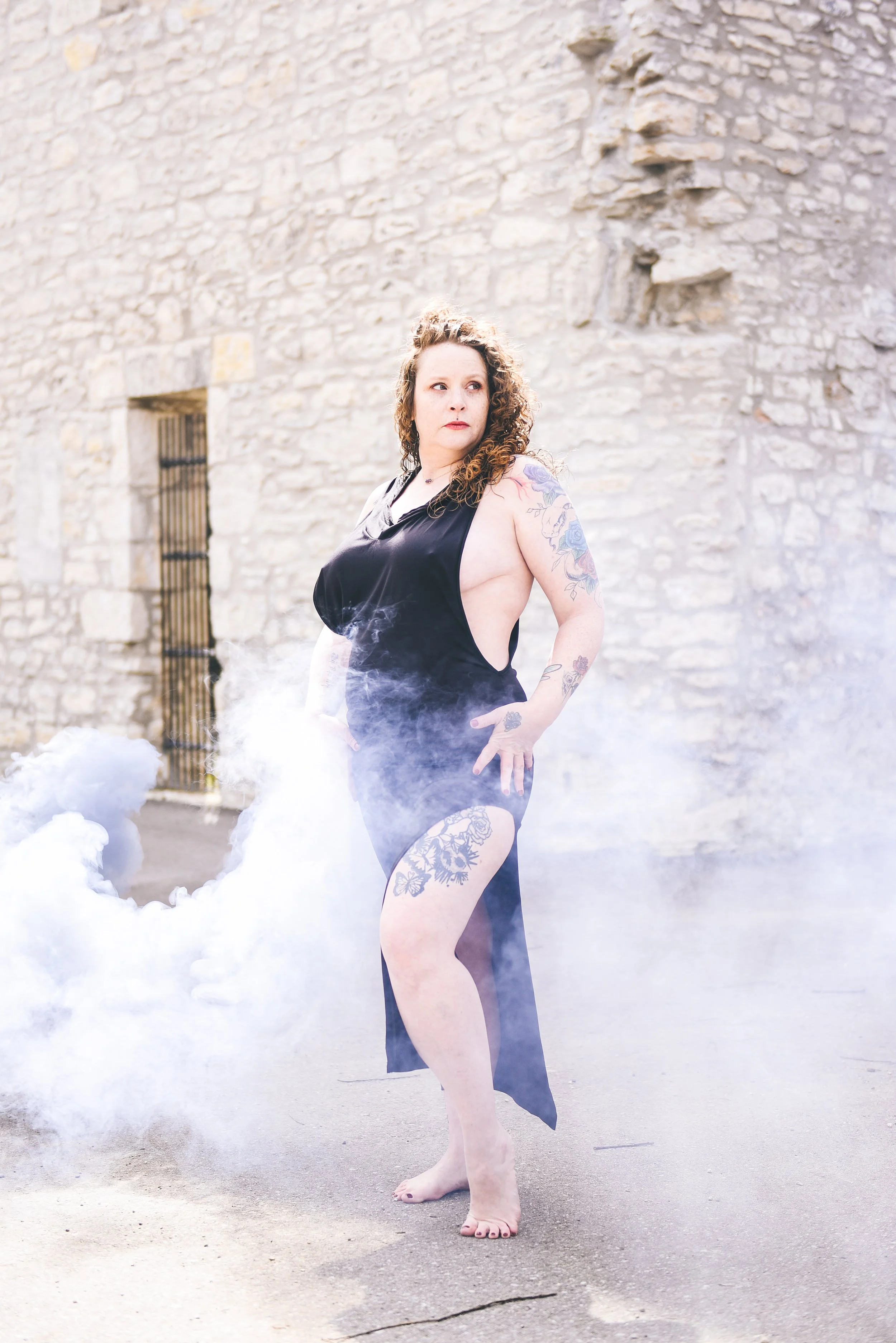 A woman with tattoos standing barefoot on pavement, wearing a black dress with open sides, surrounded by smoke or fog, with a stone wall in the background.