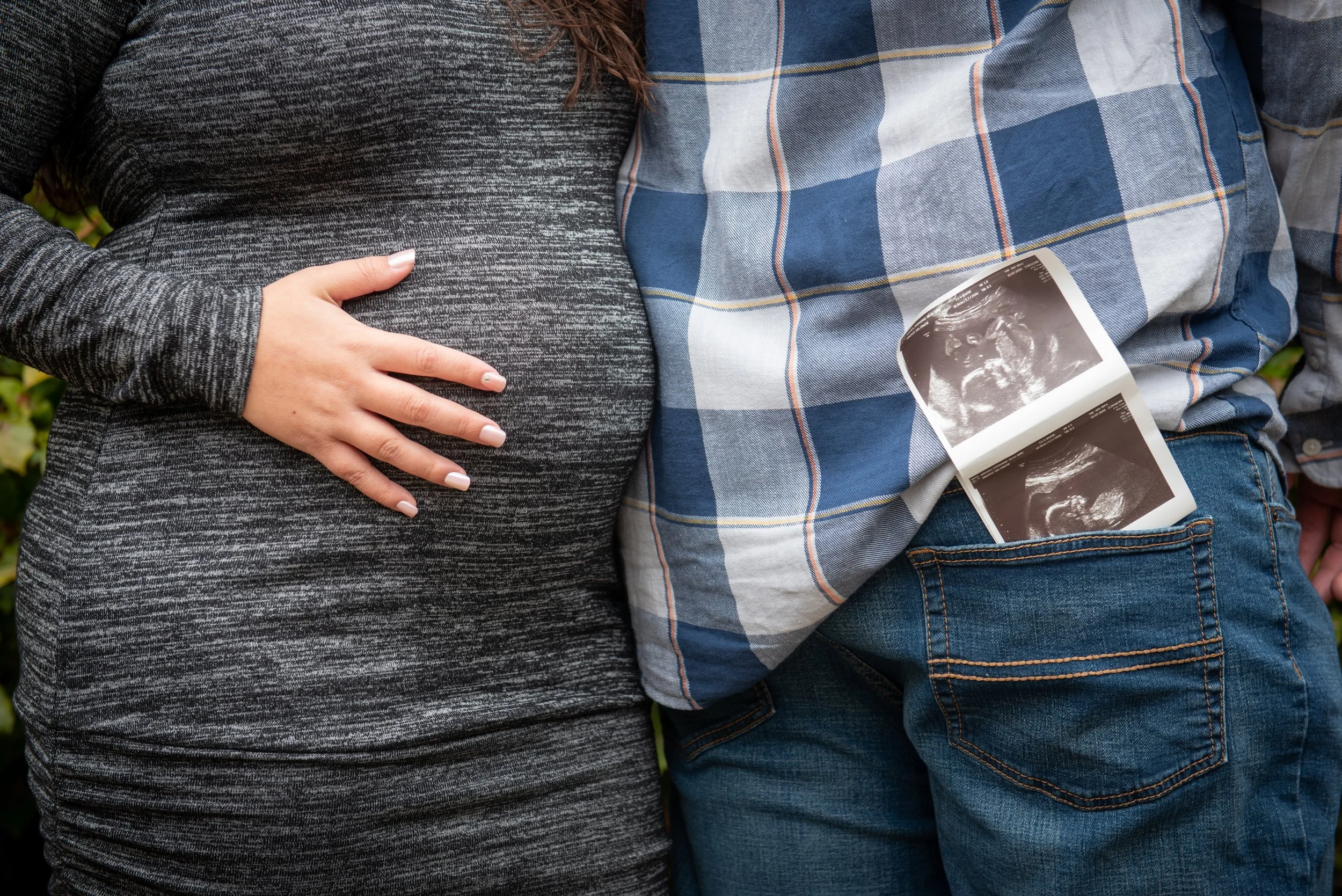 Pregnant woman touching her belly with one hand, beside a person holding ultrasound images, both standing outdoors.