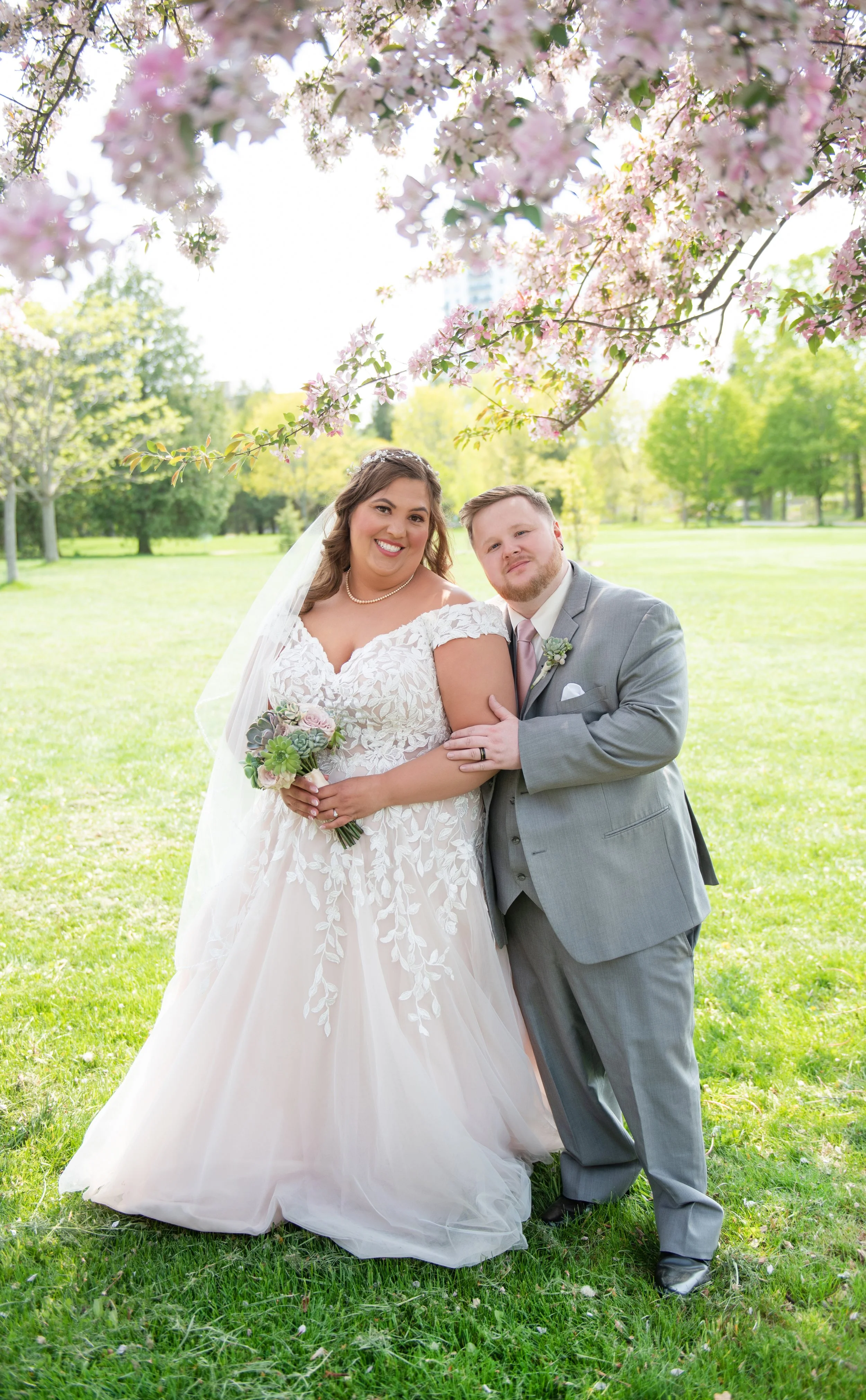 A bride in a white lace wedding dress and a groom in a light gray suit stand together under blooming cherry blossom trees in a park, smiling at the camera.
