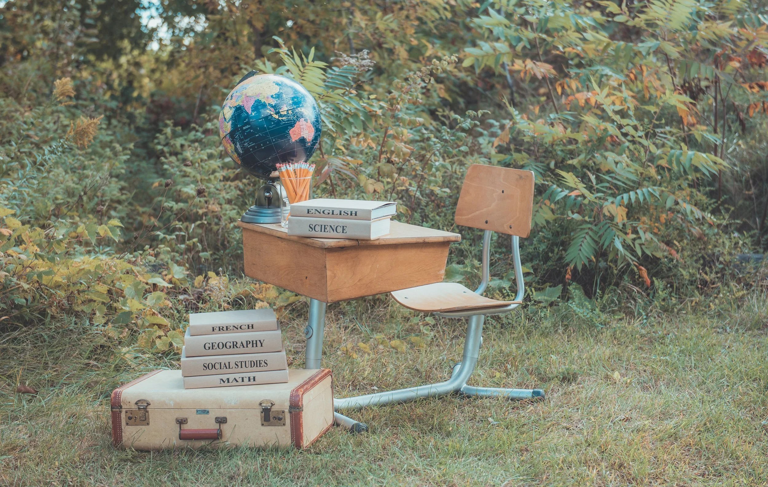 An outdoor scene with a school desk and chair placed on grass and surrounded by foliage. The desk holds a globe, a cup of pencils, and some books labeled "English" and "Science." A stack of textbooks labeled "French," "Geography," "Social Studies," and "Math" is on a vintage suitcase.