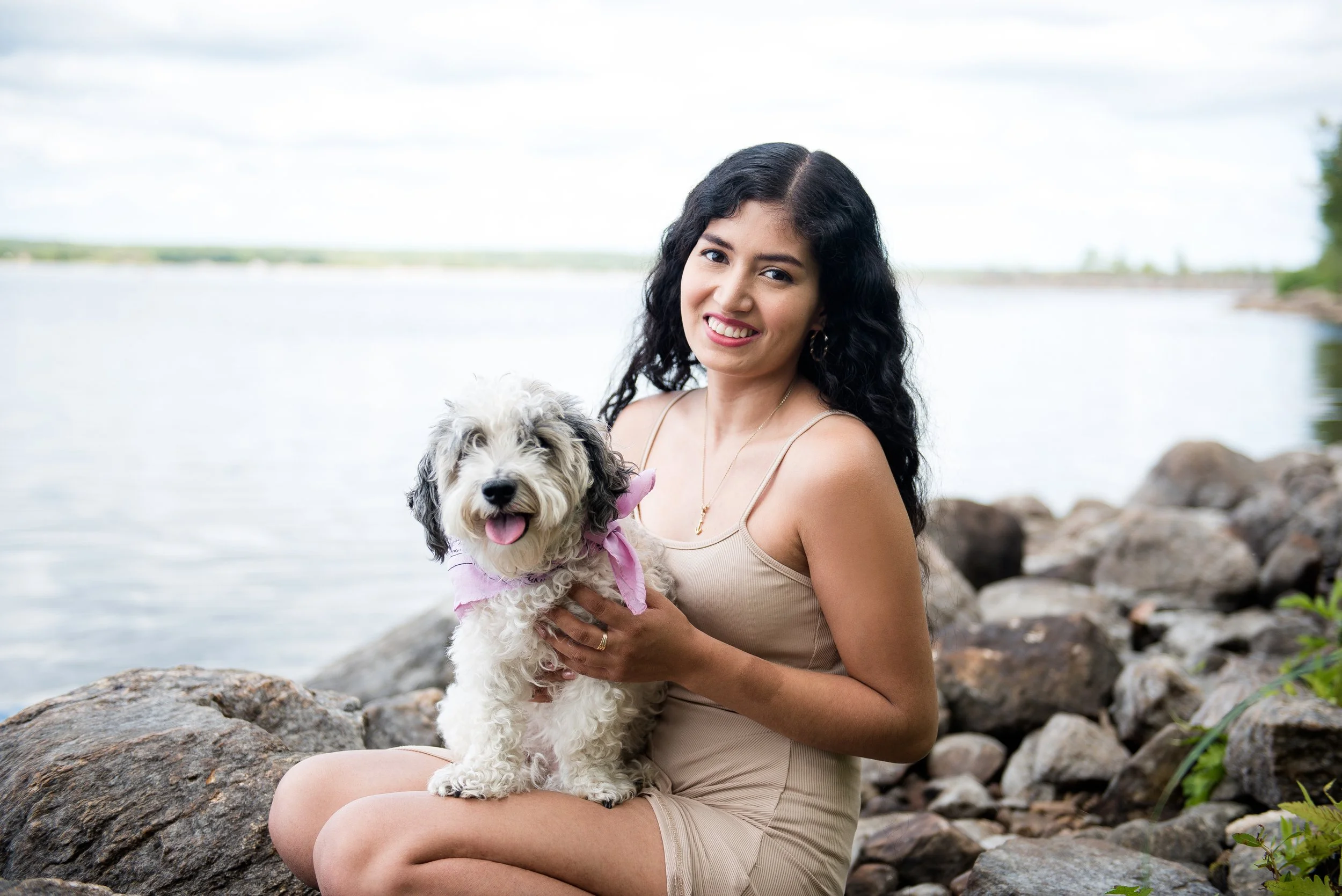 A woman with long black curly hair, wearing a beige sleeveless dress, is sitting on rocks by a body of water. She is holding a fluffy white dog with black markings, wearing a pink bandana. The woman is smiling at the camera, and the dog appears happy with its tongue out. The background shows water and a cloudy sky.