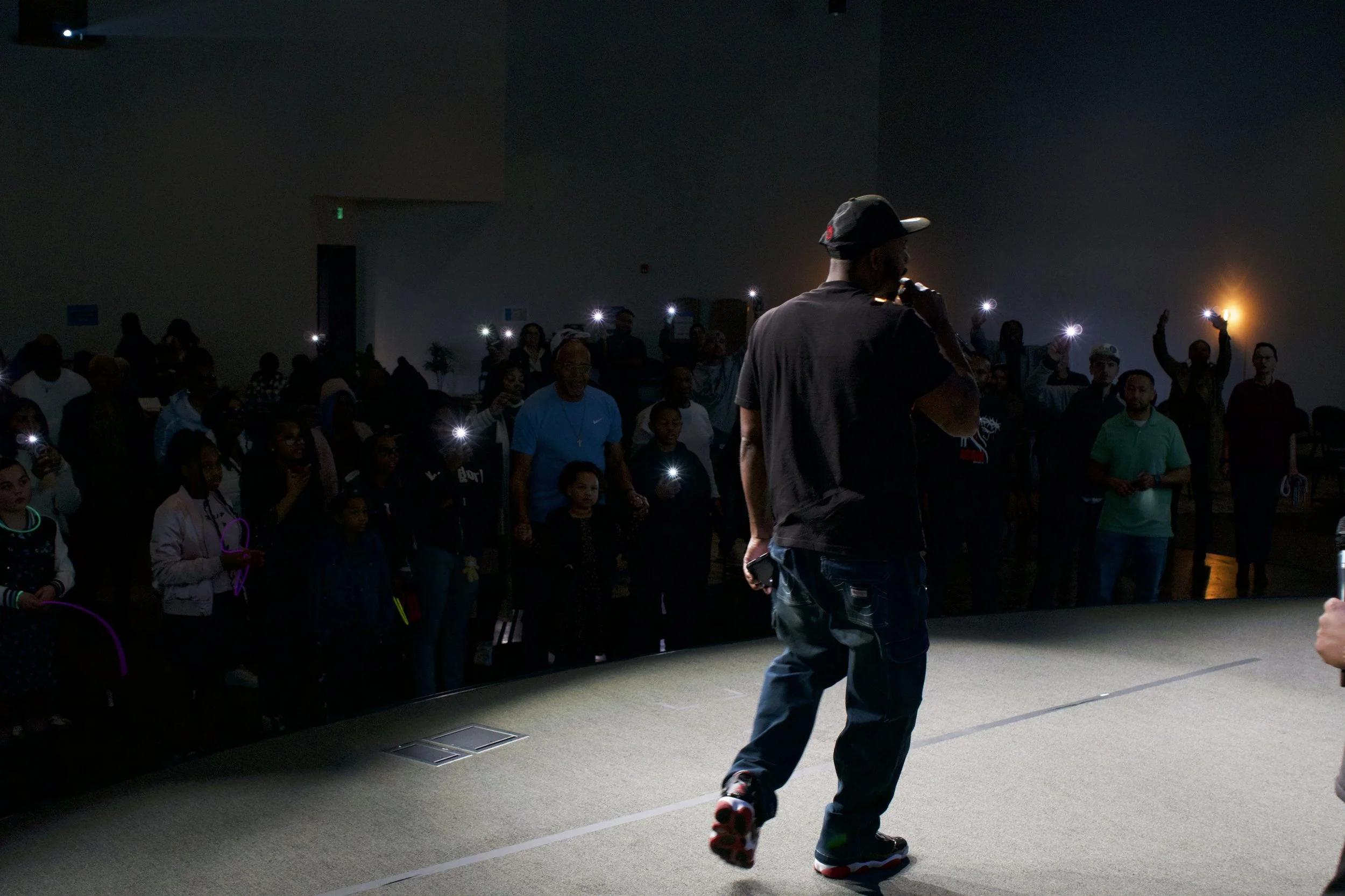 A man in a black t-shirt, jeans, and sneakers standing on a stage, holding a microphone, while a crowd of people in a dark room behind him is taking photos with their phones. The crowd includes adults and children, and the room is dimly lit.