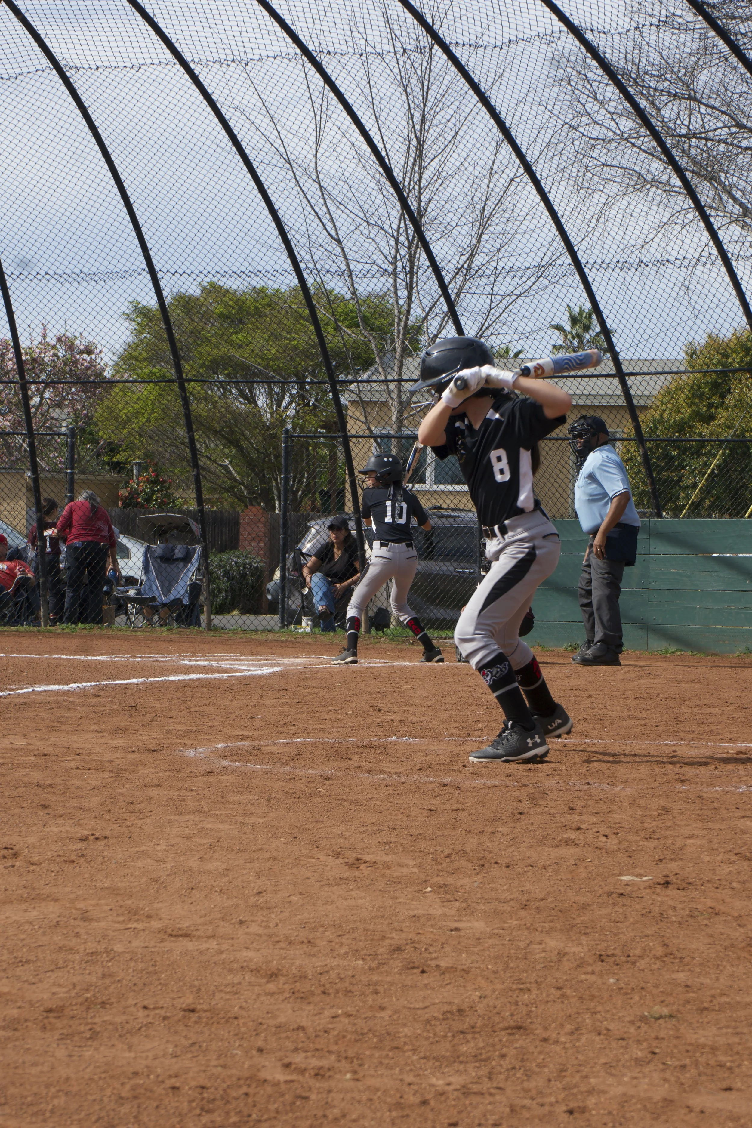 Girls playing softball on a dirt field with a chain-link fence and spectators in the background.