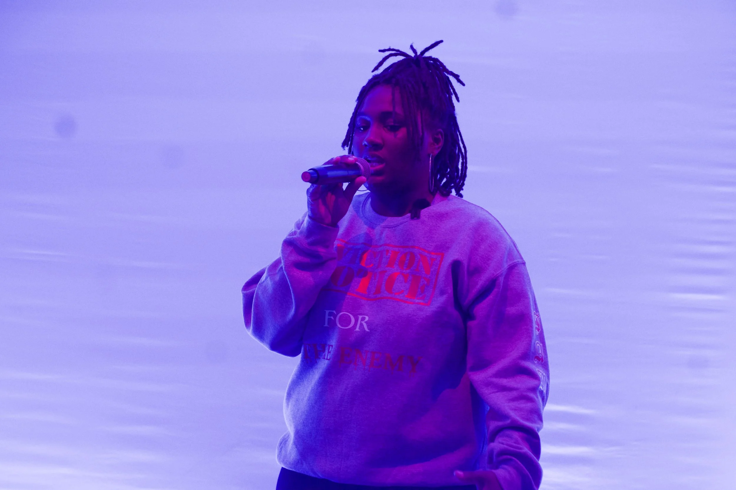 A woman with dreadlocks holding a microphone and singing or speaking against a plain background.