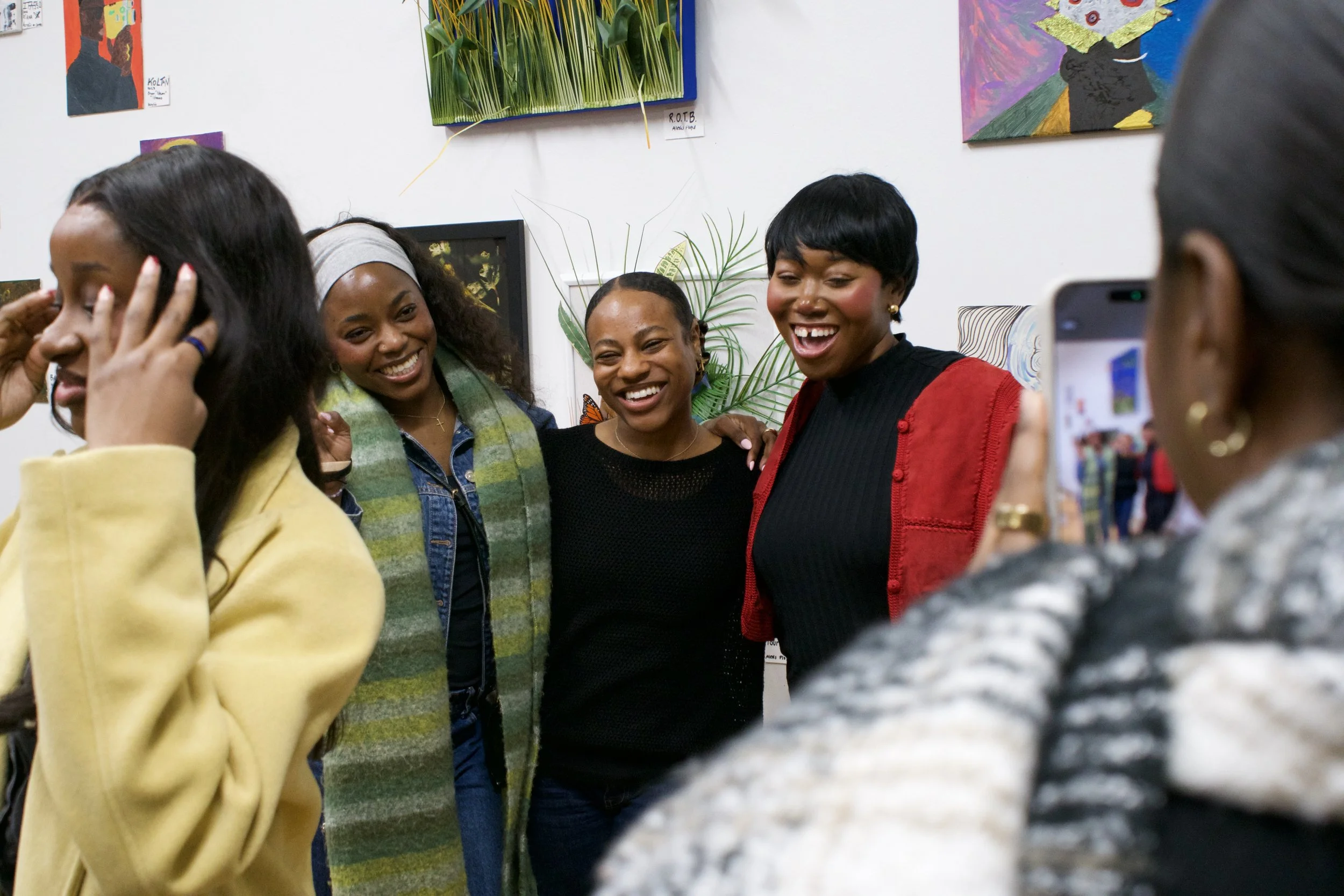Four women are gathered together, smiling and posing for a photo in an art gallery or exhibition space, with paintings and artwork on the wall behind them.