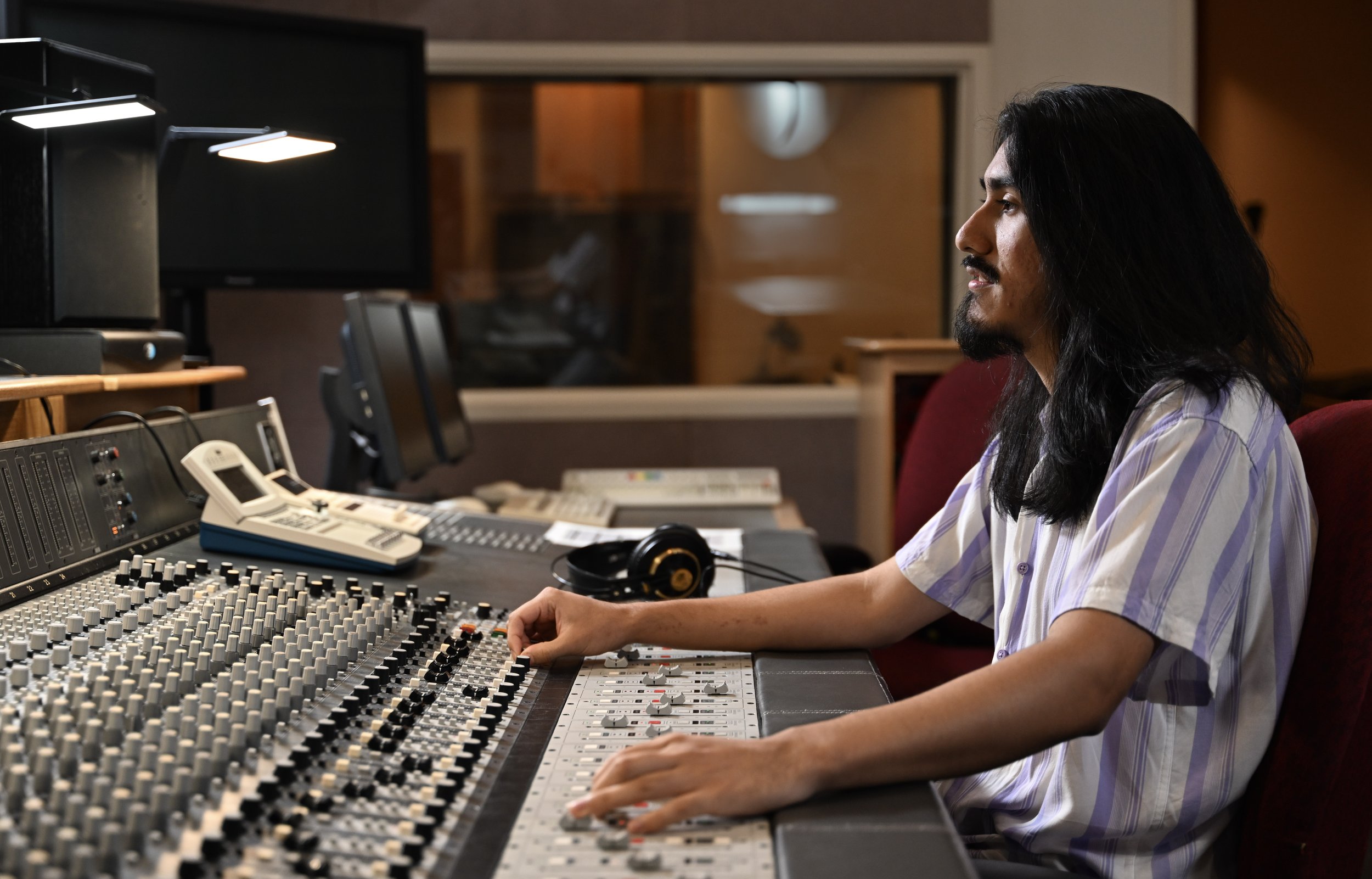 A man with long black hair and a beard working at a recording studio mixing console, wearing a striped shirt with headphones on the desk, in a professional sound recording environment.