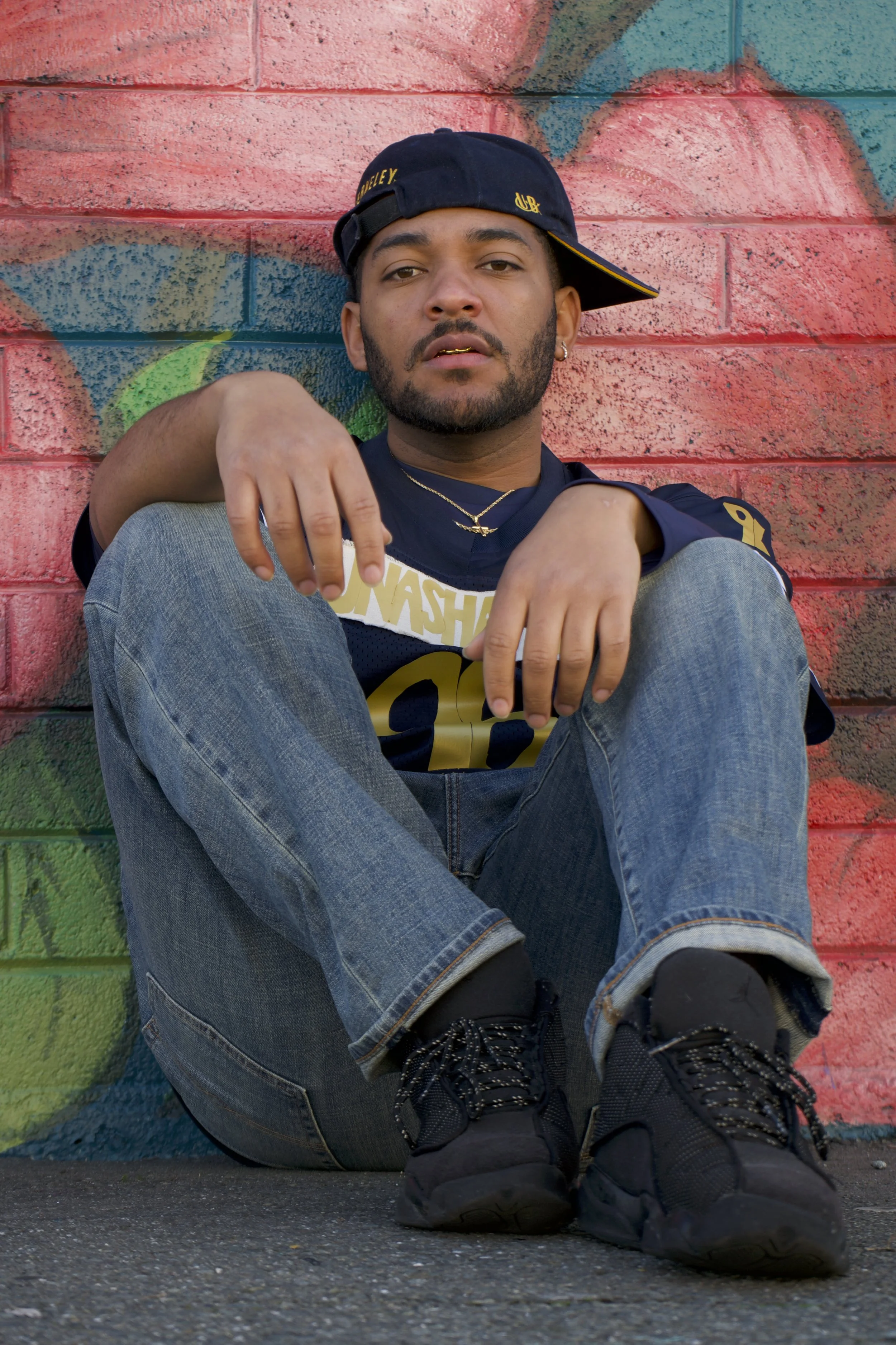 A young man sitting on the ground with knees up, wearing a black cap, black sneakers, jeans, and a sports jersey, against a colorful graffiti wall.