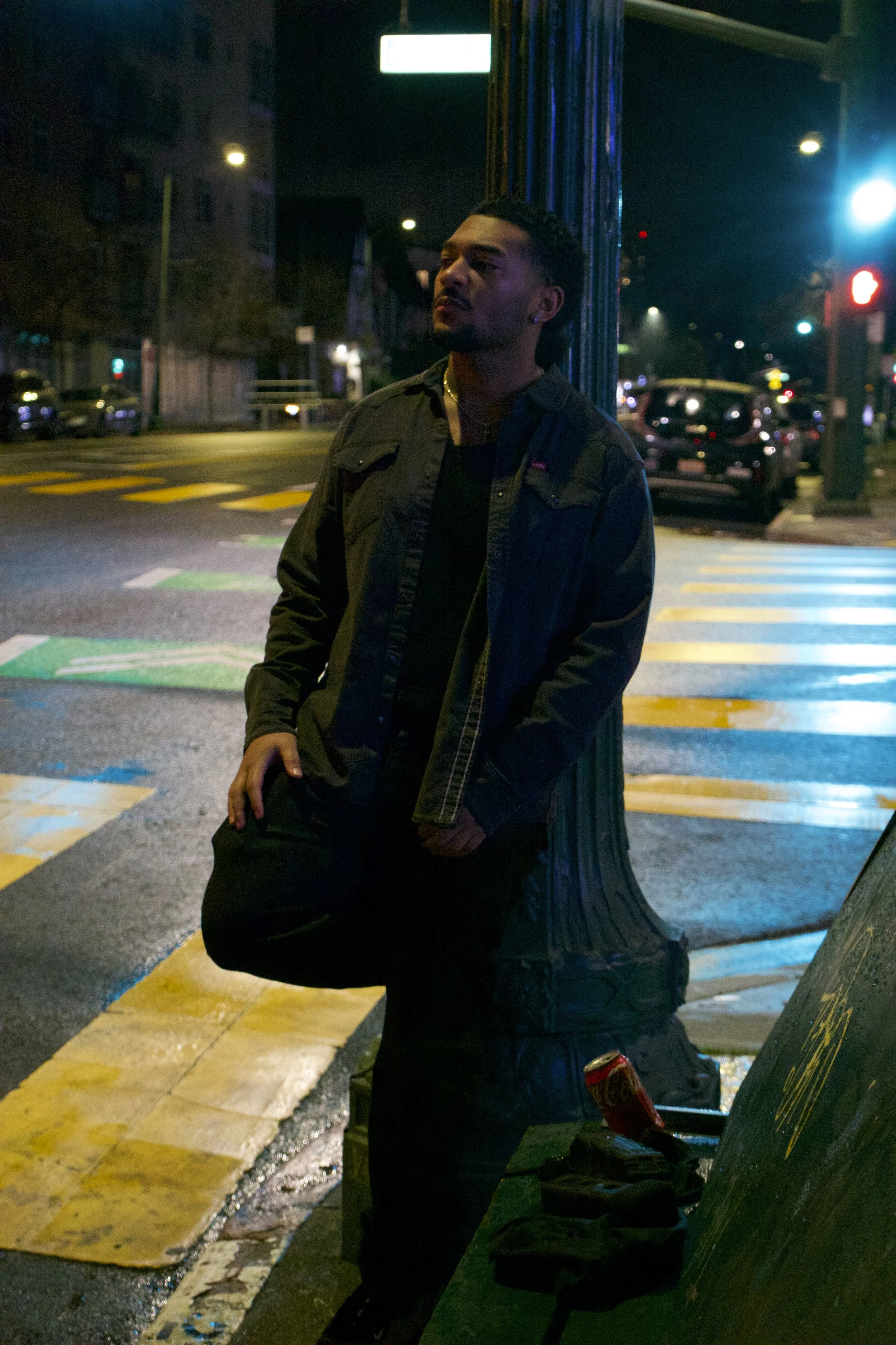 A man stands on a city street at night, leaning against a utility pole, with cars parked behind him and colorful street lighting.