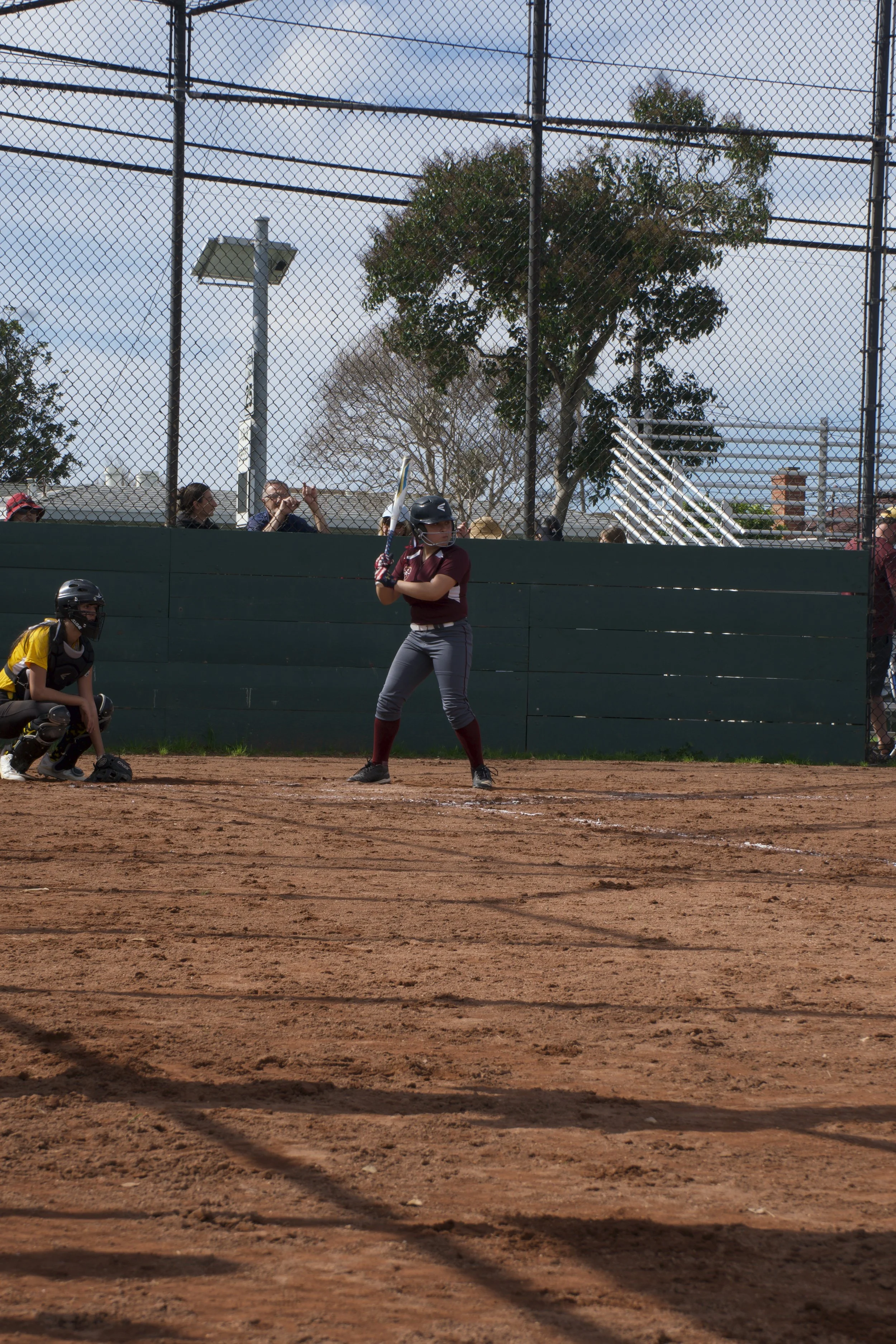 A girl in a maroon shirt and gray pants at bat on a baseball or softball field with dirt infield, holding a bat ready to hit. A catcher in a black and yellow uniform with a mask is crouched behind her, and spectators are behind a green fence, with tr