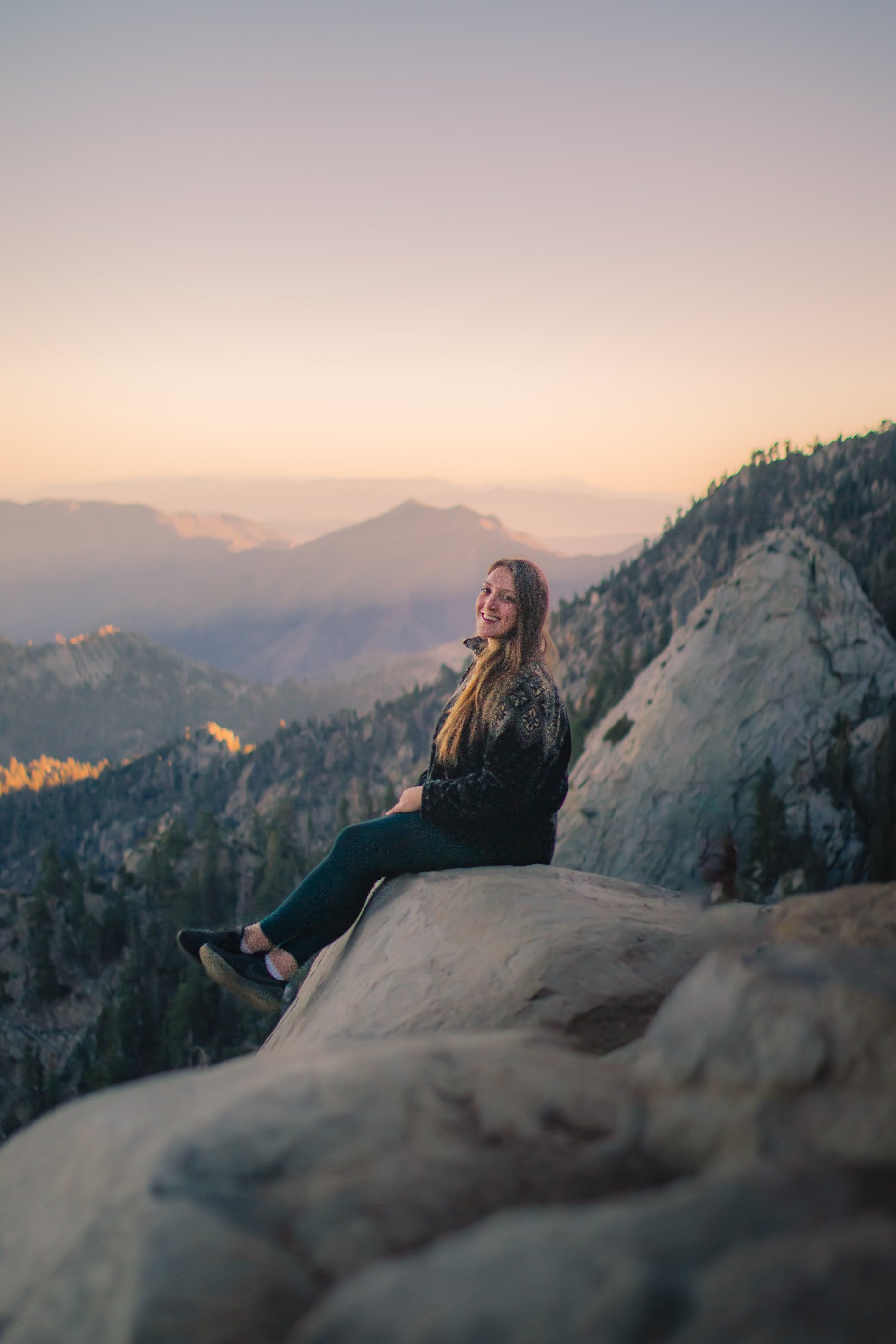 A woman sitting on a large rock formation in a mountainous landscape during sunset, smiling and looking at the camera.