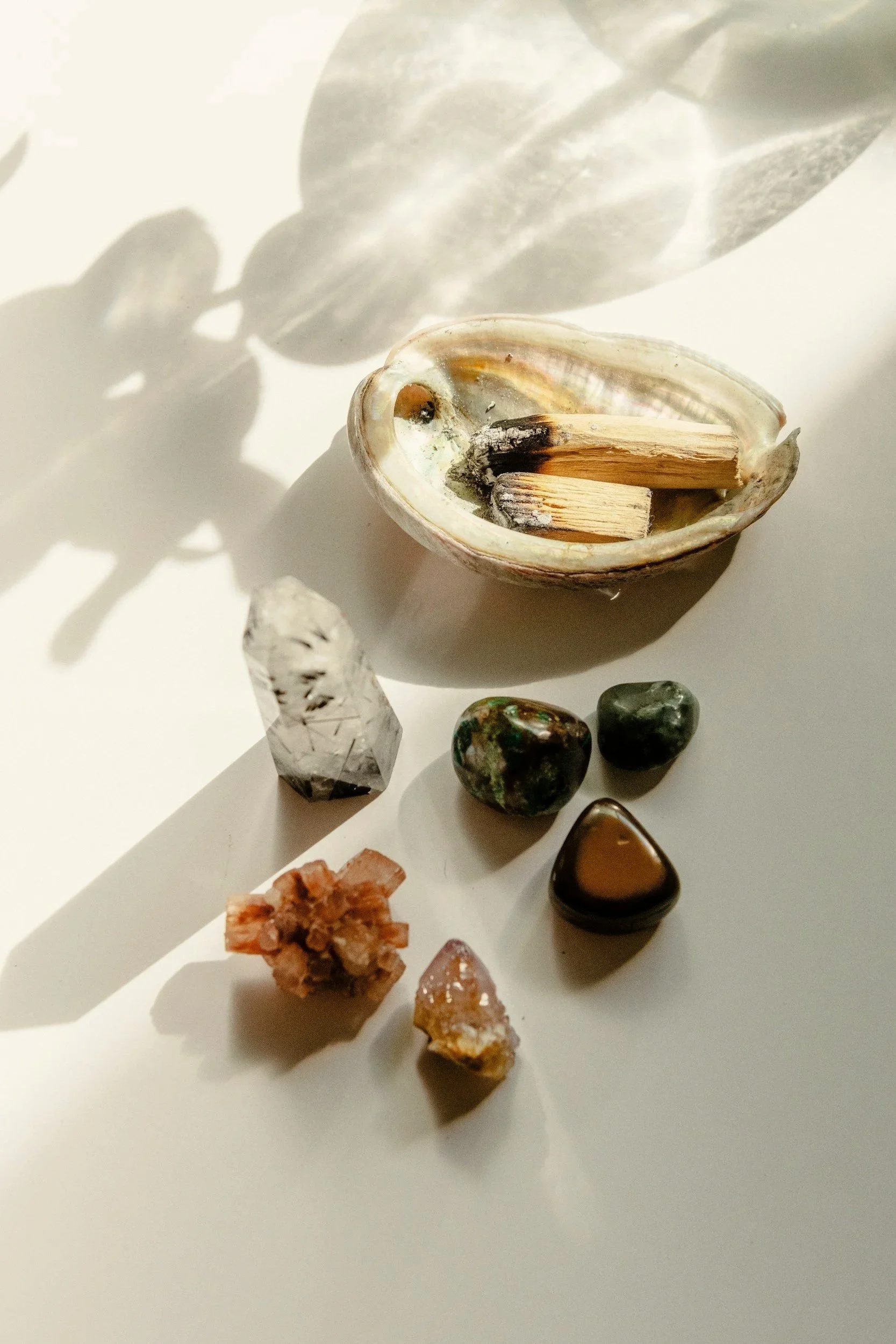 An open seashell with two matchsticks inside, various polished stones, and a mineral cluster resting on a white surface with shadows cast by sunlight.