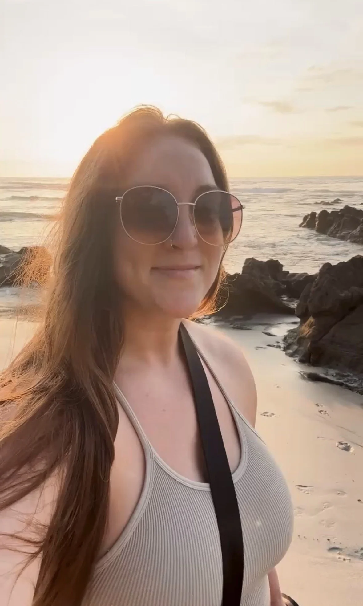 A woman with long brown hair wearing sunglasses and a beige tank top on a beach during sunset.