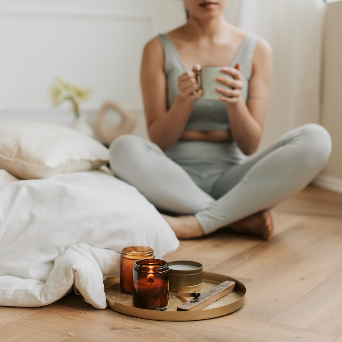 A woman sitting cross-legged on the floor, holding a mug, with candles and a tray in front of her, beside a bed with white bedding and a pillow.
