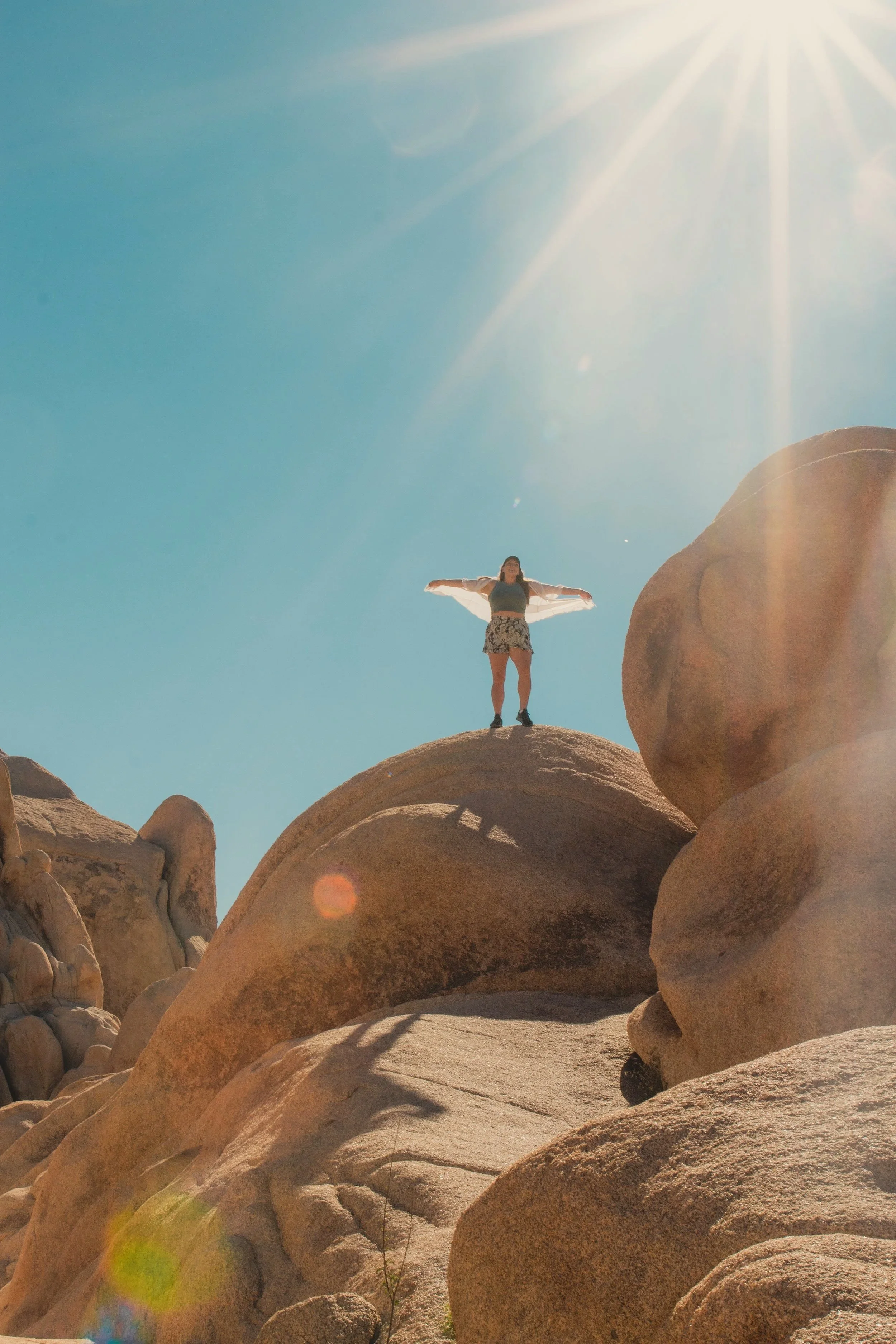 A person standing on a large rock formation in a desert area with a clear blue sky and the sun shining brightly overhead.
