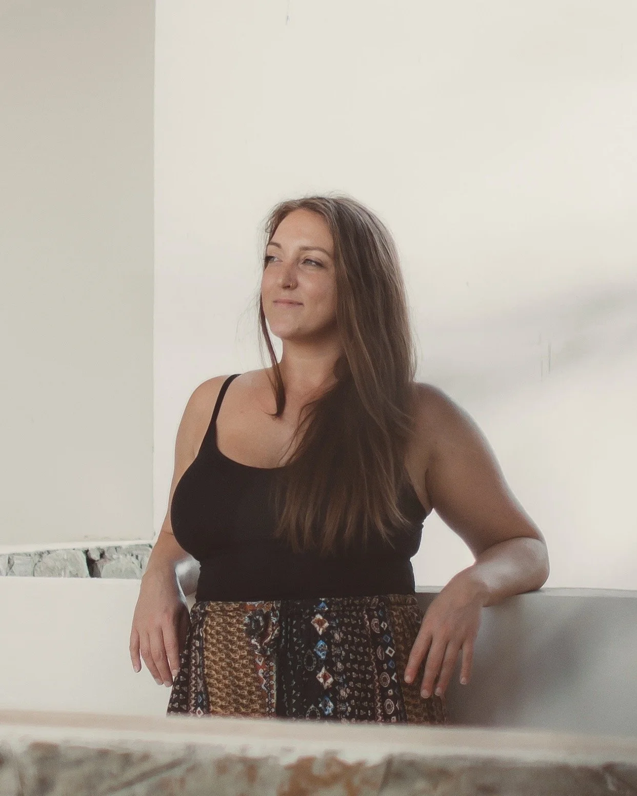 A woman with long brown hair wearing a black tank top and patterned skirt. She is standing indoors, leaning on a white railing, with a slight smile and looking to the side.