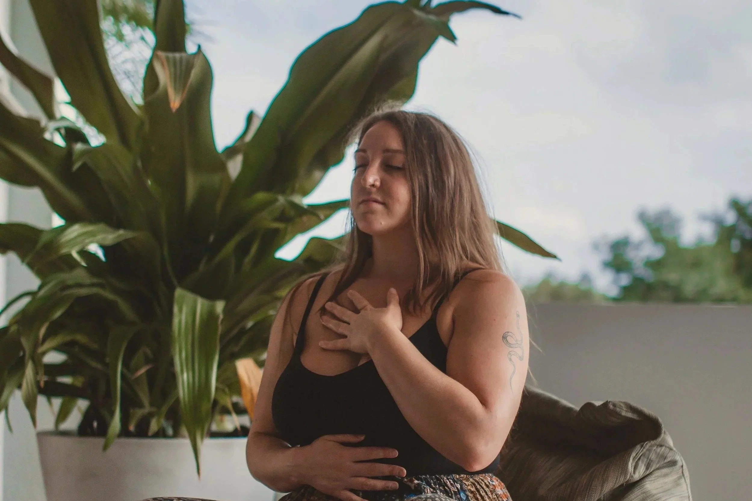 A woman with long brown hair, wearing a black tank top, sitting on a couch with her eyes closed, hand on her chest, in a peaceful moment next to a large green houseplant, with a window showing trees outside.