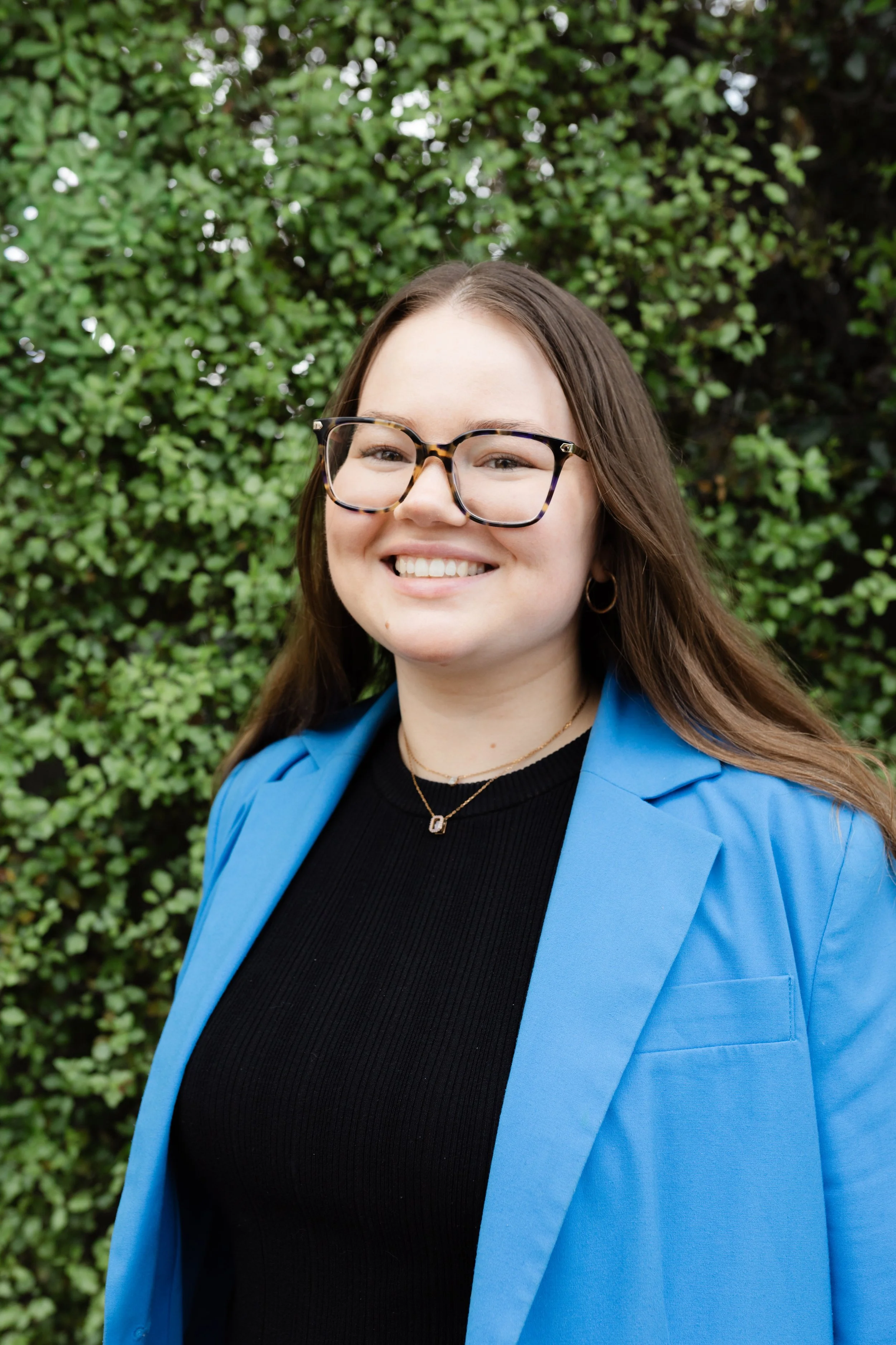 A woman with long brown hair, wearing tortoiseshell glasses, a black top, a blue blazer, and gold earrings, smiling outdoors in front of green leafy bushes.