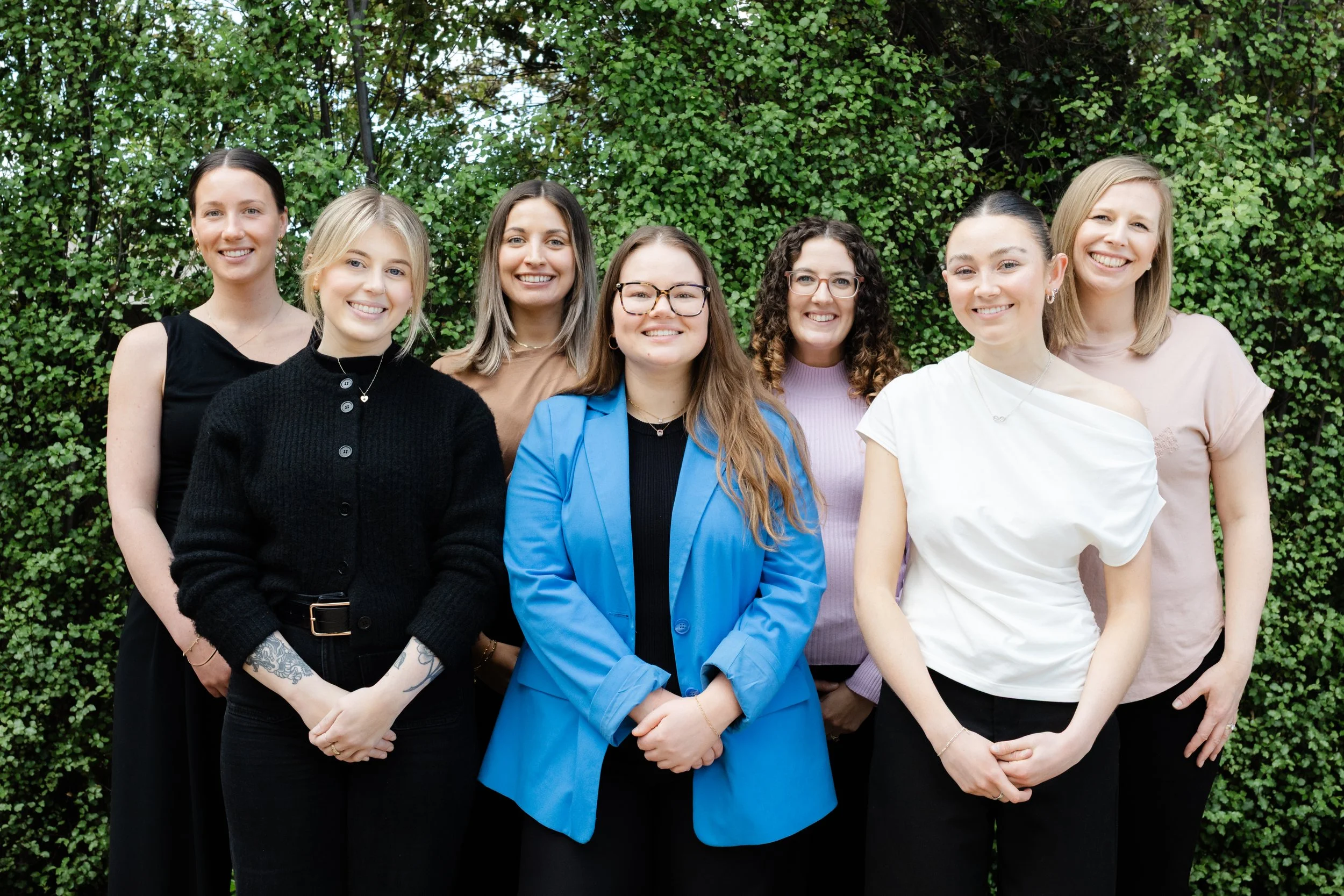 Group of seven women smiling outdoors standing in front of green leafy hedge.