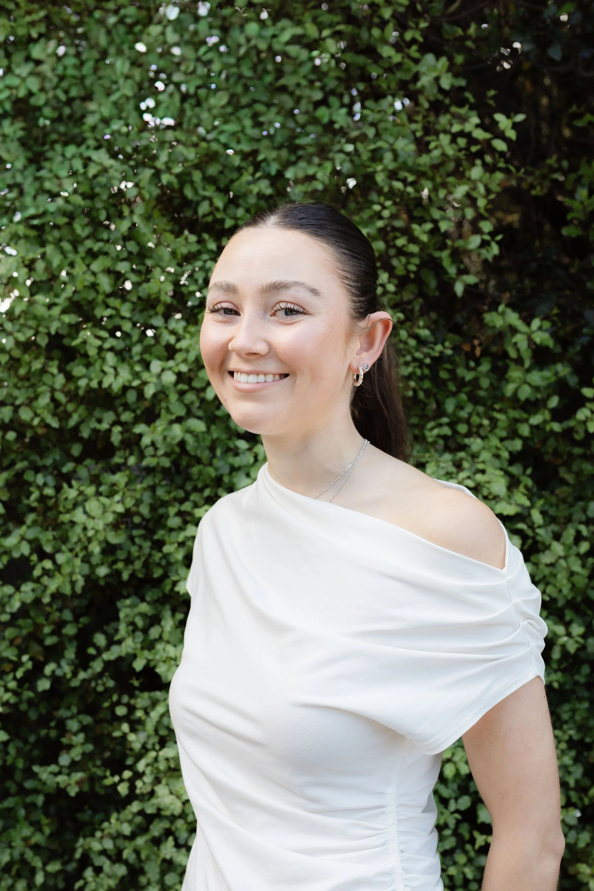 A young woman with dark hair tied back, smiling, wearing a white off-shoulder top, earrings, and standing in front of a green leafy background.