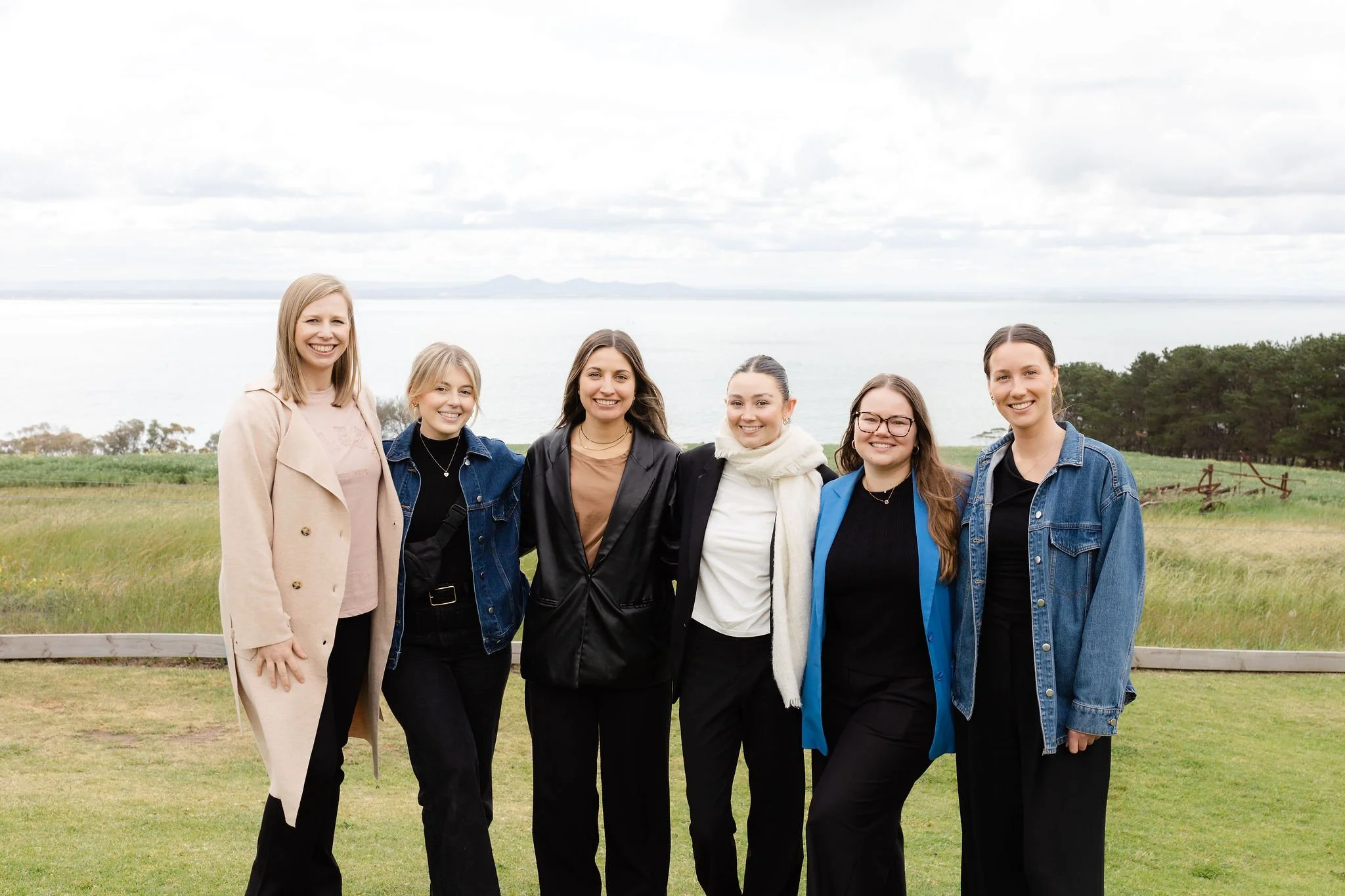 A group of six women standing outdoors on grass near water with hills and trees in the background, smiling at the camera.