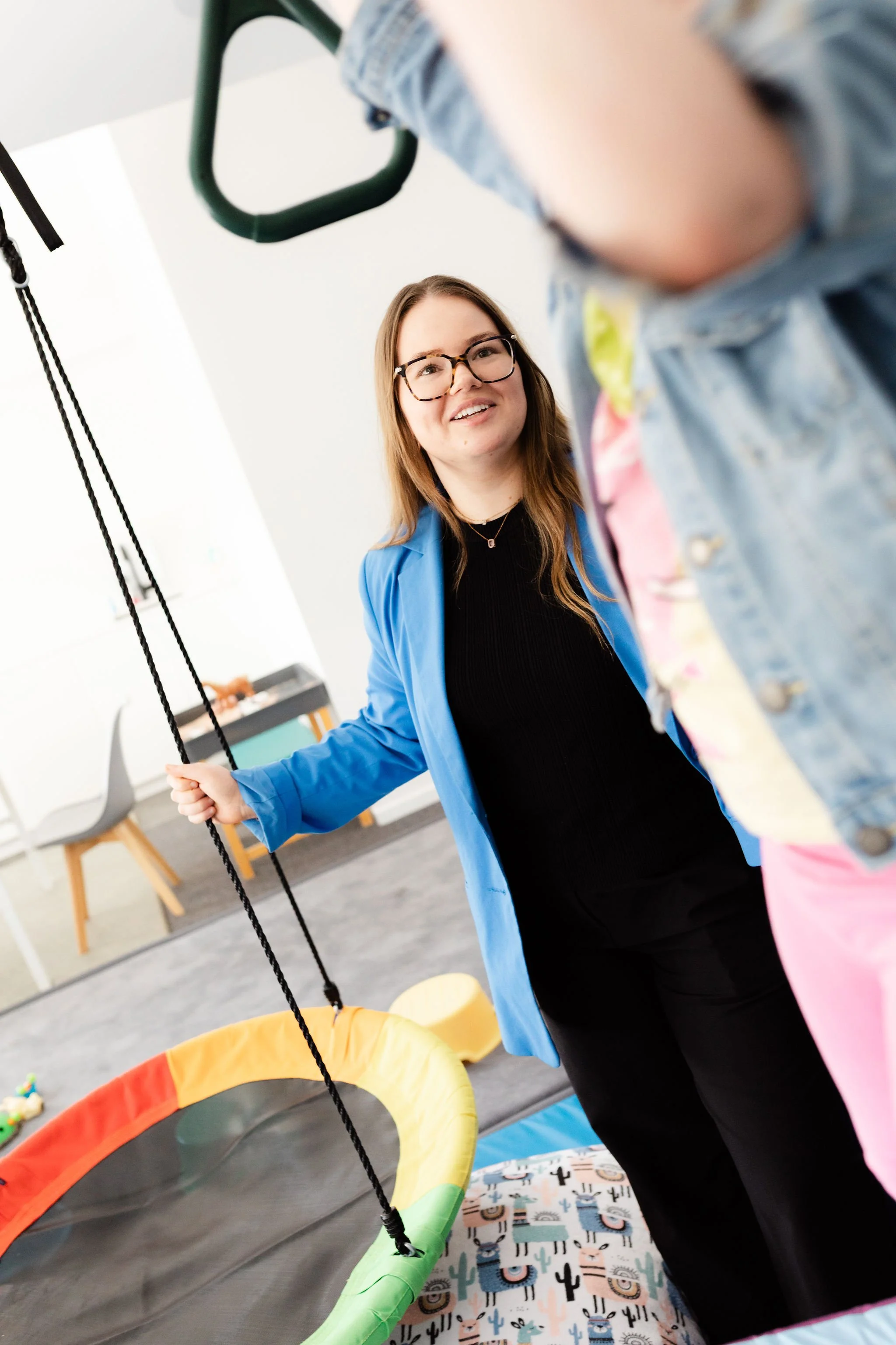 Woman with glasses and long hair smiling, wearing a blue jacket, playing on a children's indoor play area with a rainbow-colored bounce house and a patterned mat with llamas and cacti.