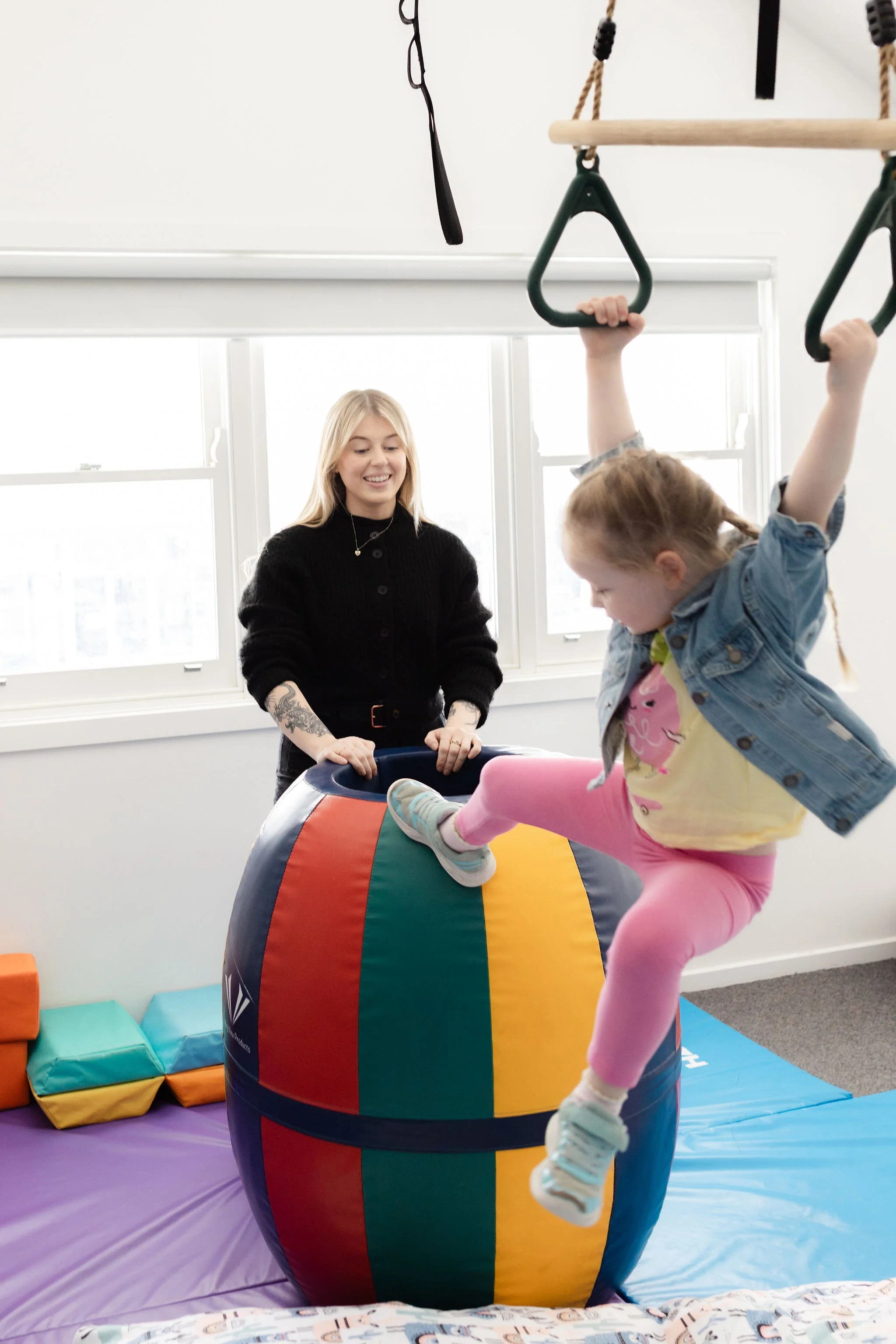 A young girl bouncing on a colorful inflatable ball in a playroom with a woman supervising, with gymnastic rings hanging from the ceiling.