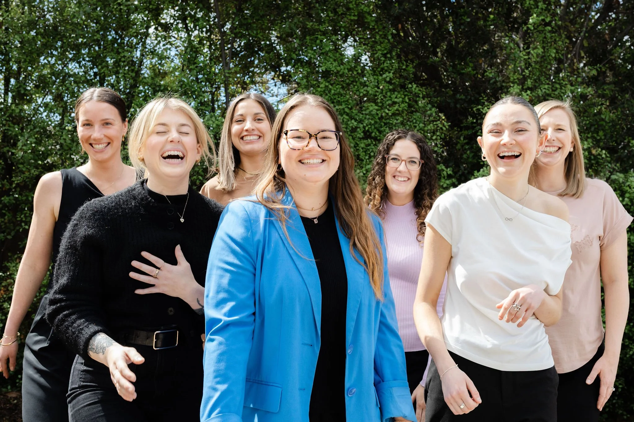 A group of seven women smiling and laughing outdoors, standing in front of green trees. They are dressed casually, with some wearing glasses and colorful tops or jackets.