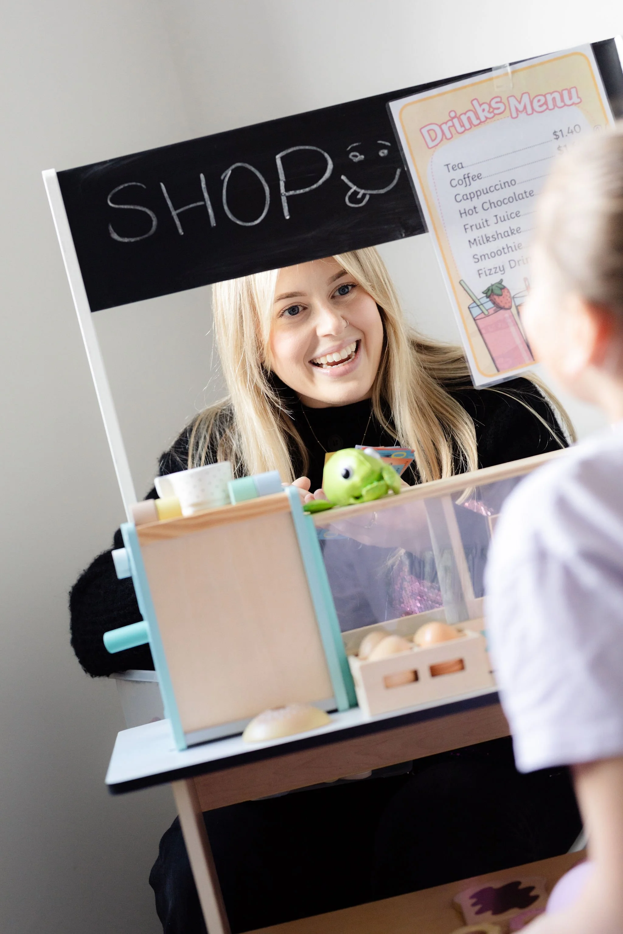 A smiling woman working at a small booth with a blackboard that has 'SHOP & : )' written on it and a nearby drinks menu. The booth has a pink and orange color theme, with various drinks and small items on display.