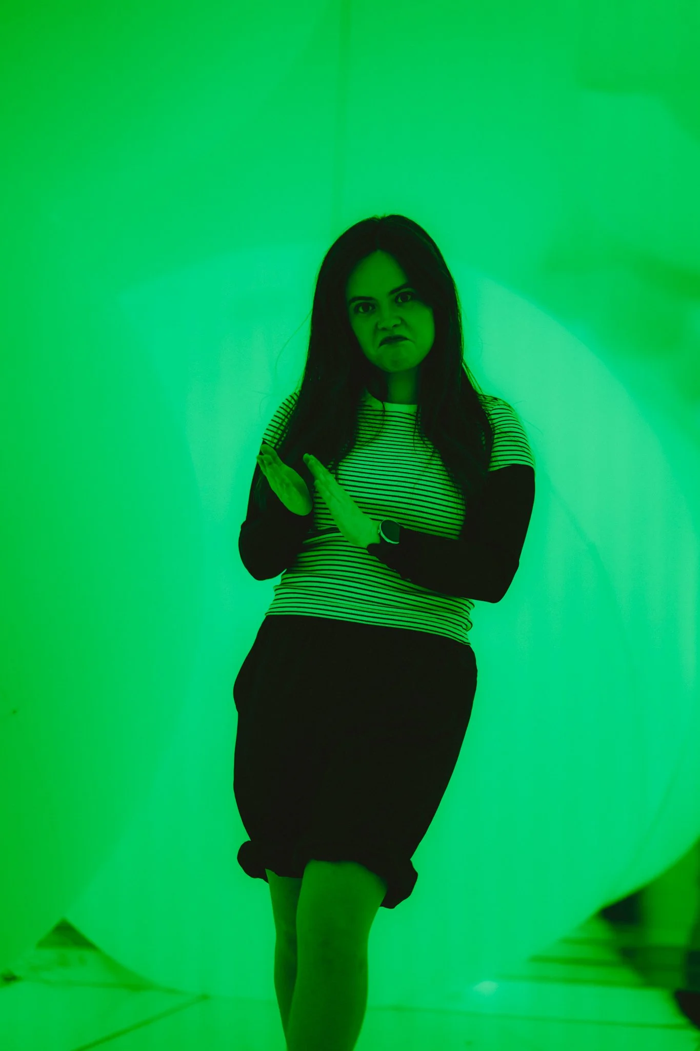 A woman with long dark hair wearing a striped shirt and black skirt making a disapproving expression with green lighting.
