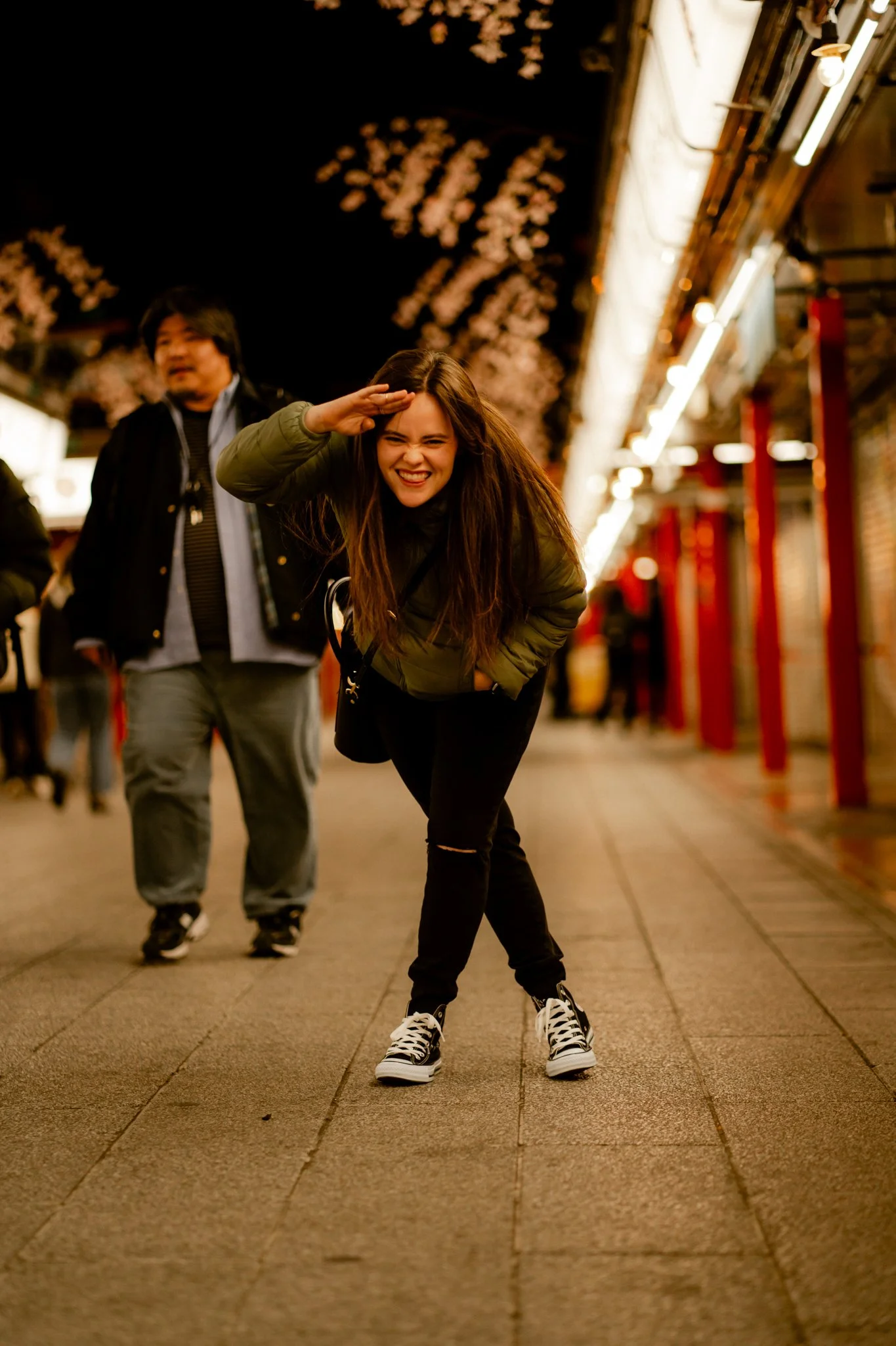 A woman laughing and saluting on a city sidewalk at night, with a man standing behind her, under string lights and cherry blossom decorations.