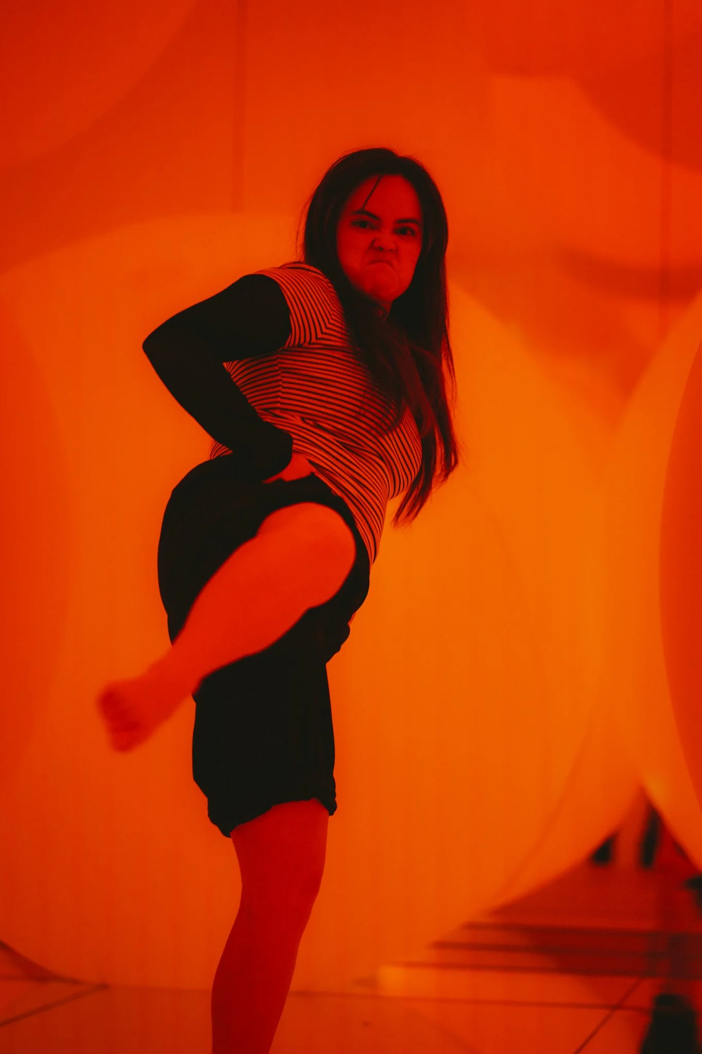 A woman with long dark hair making a fierce face, standing against an orange background with circular designs, wearing a striped shirt with black sleeves and black shorts.